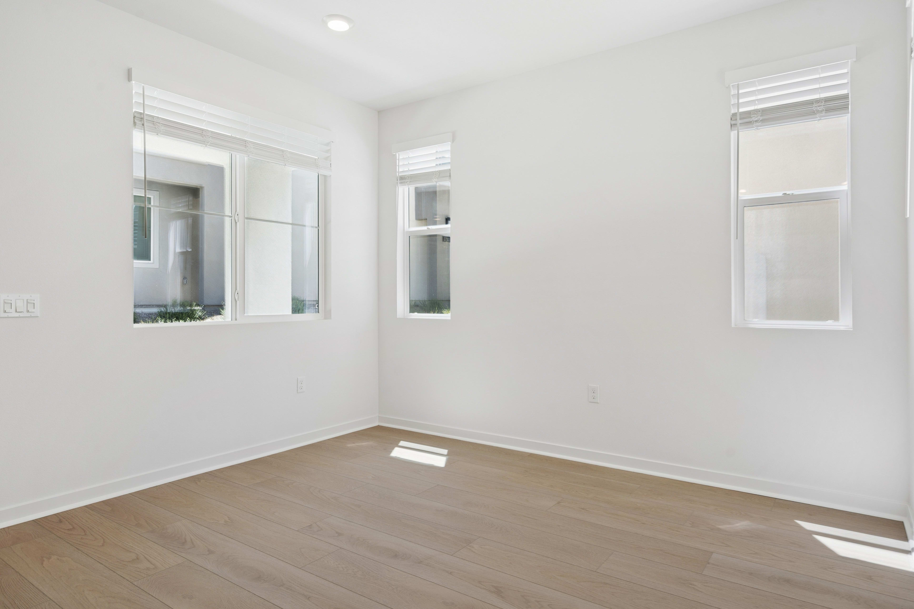 A bright, minimalist room with white walls, wooden flooring, and large windows allowing natural light to fill the space.