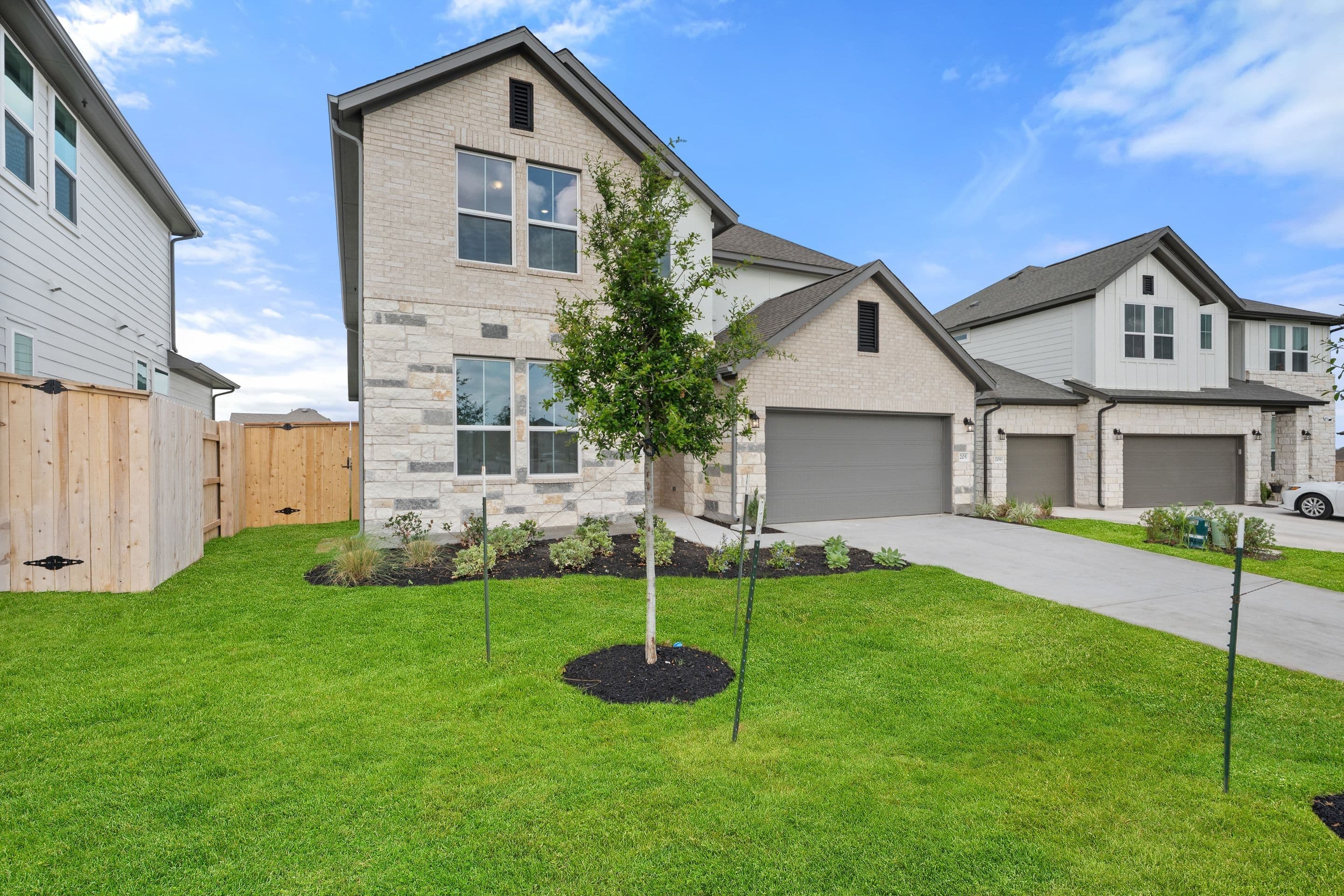 The image depicts a newly constructed residential neighborhood with several modern, two-story houses featuring gray siding and large windows. The foreground showcases a well-manicured lawn with a young tree, creating a pleasant and inviting outdoor space.