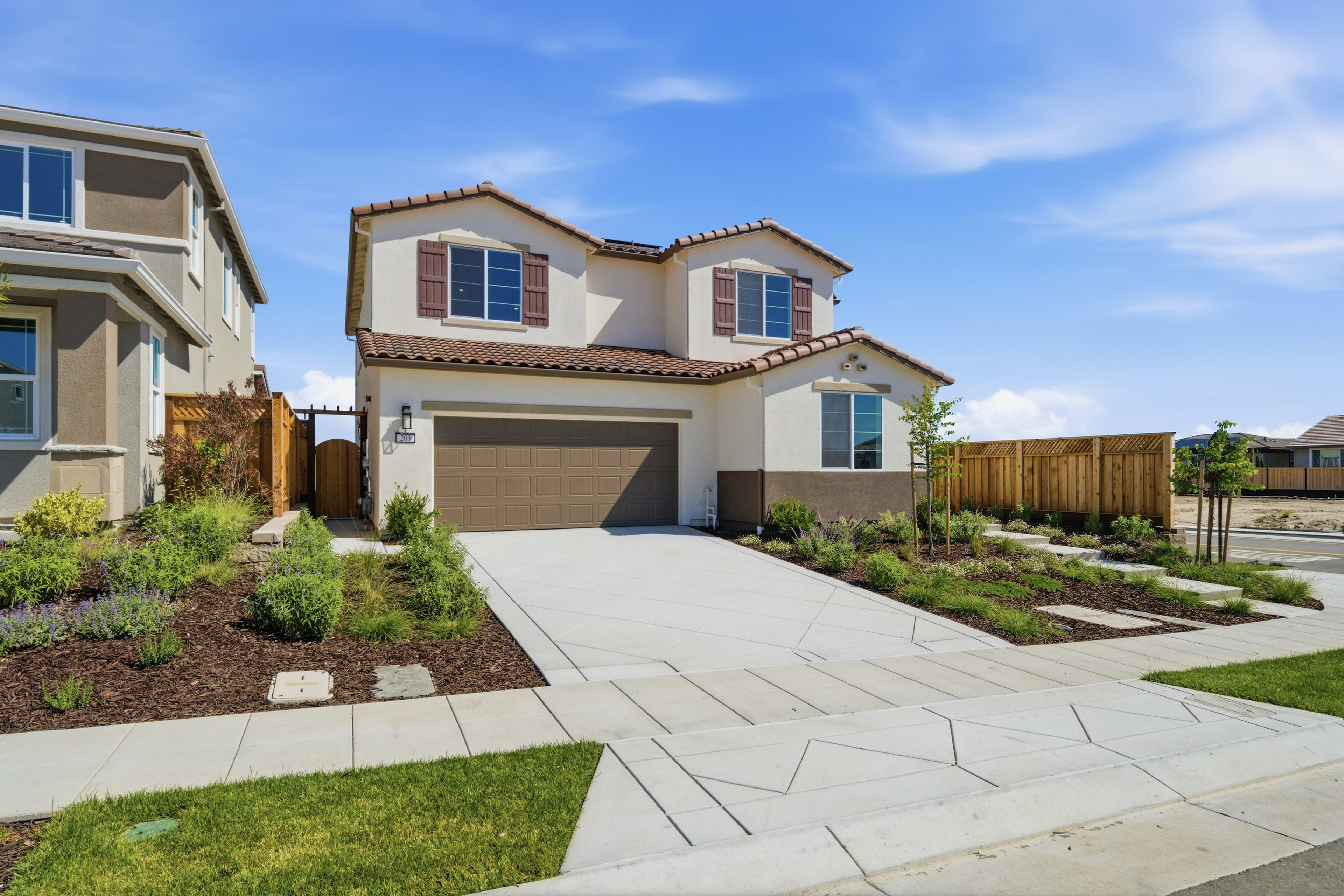 A two-story residential house with a well-manicured lawn and landscaping, set against a clear blue sky with scattered clouds.