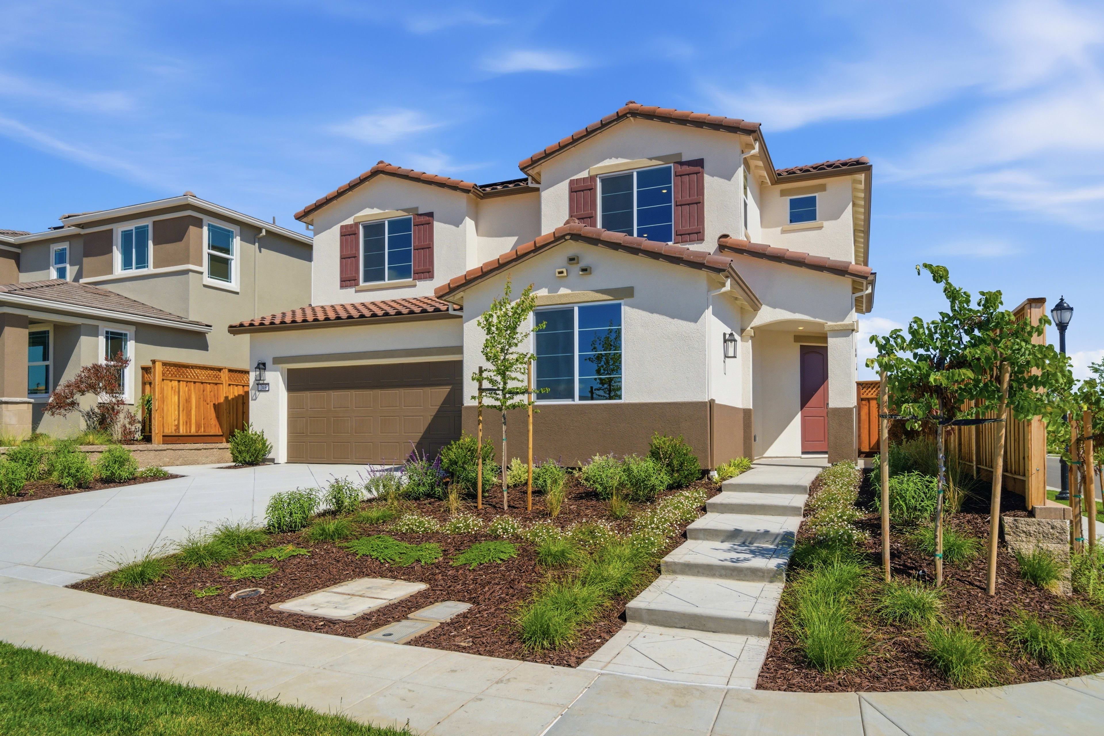 A two-story Mediterranean-style house with a tiled roof, surrounded by a well-landscaped yard featuring a paved walkway, lush greenery, and a clear blue sky in the background.