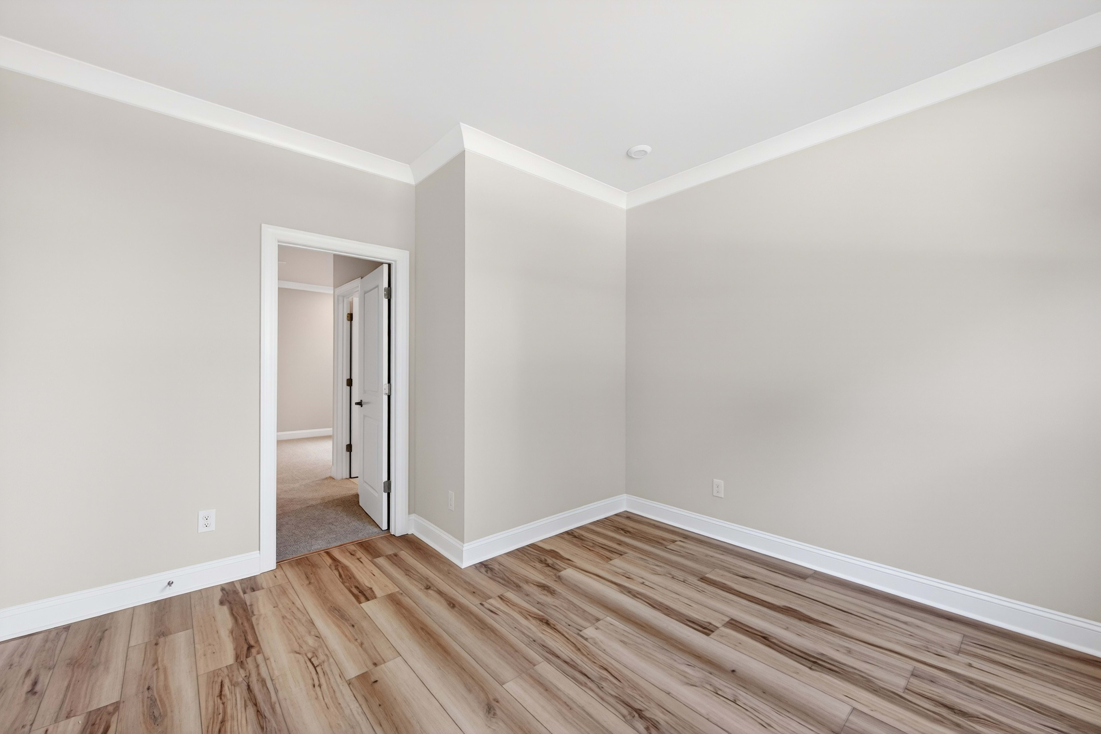 A spacious, well-lit room with white walls and a wooden floor, featuring an open doorway leading to another room.