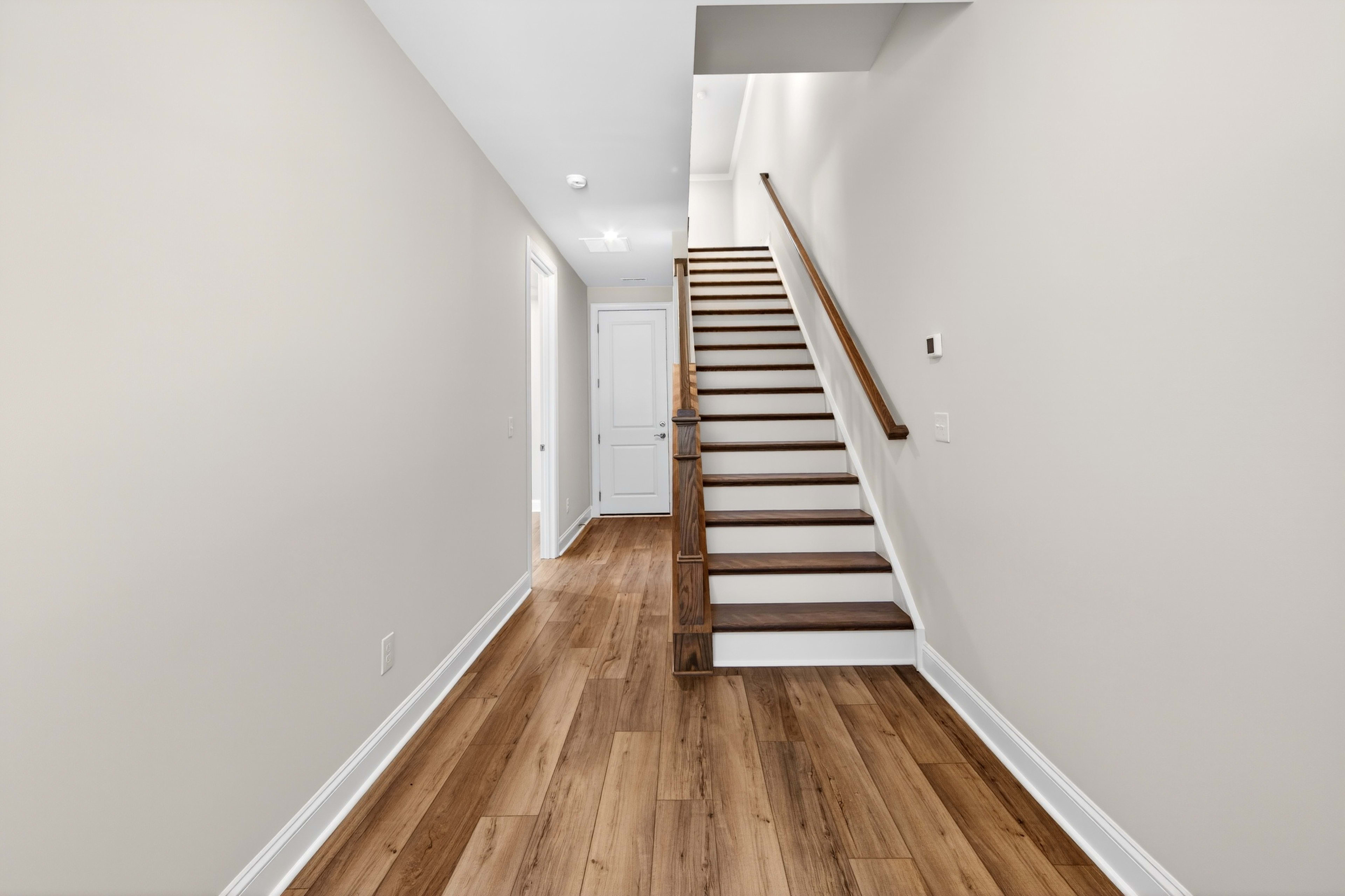 A long, narrow hallway with a wooden staircase leading up, and a hardwood floor running the length of the space.