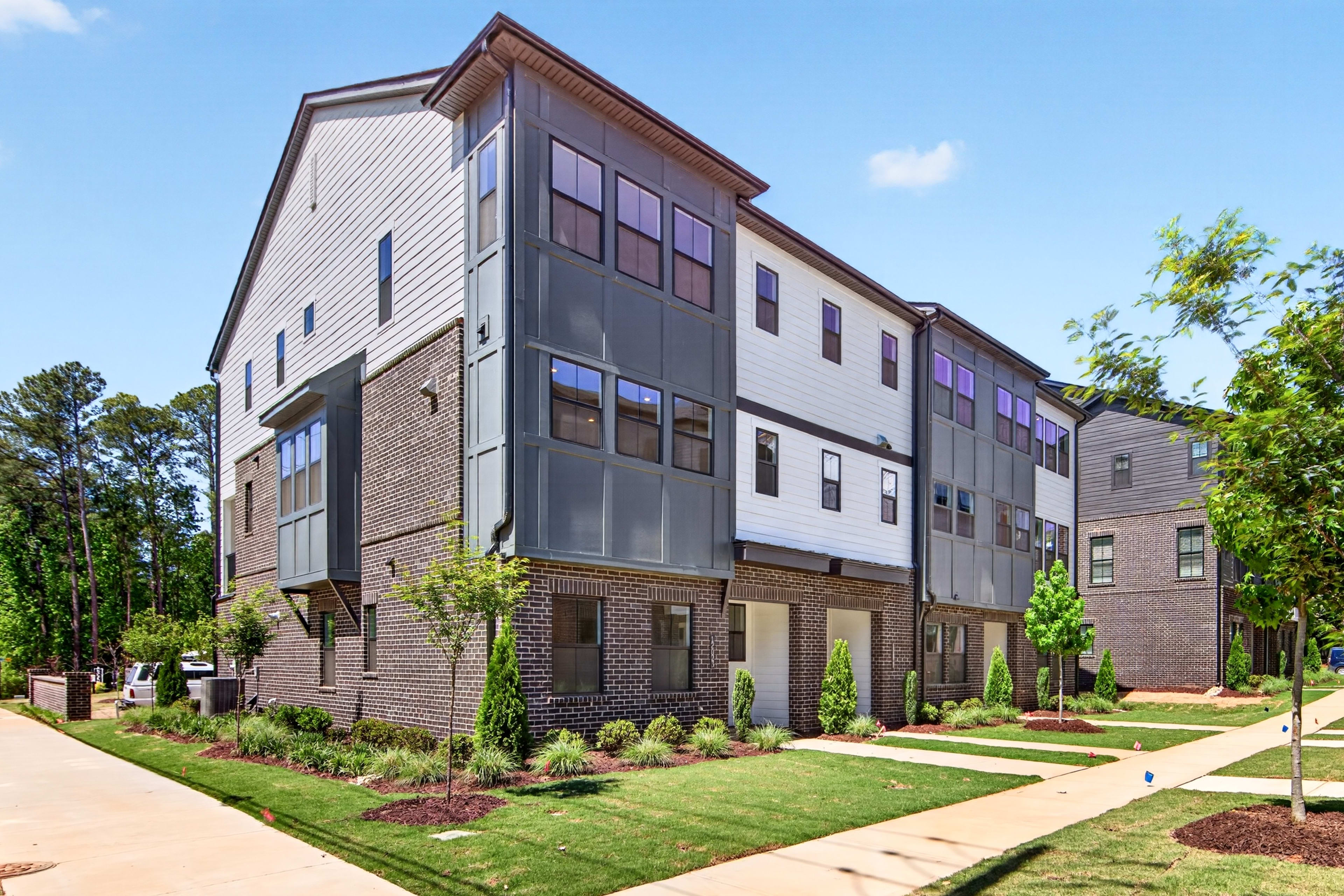 A multi-story residential building with a brick and siding exterior, surrounded by a well-manicured lawn and landscaping, set against a backdrop of trees and a clear blue sky.