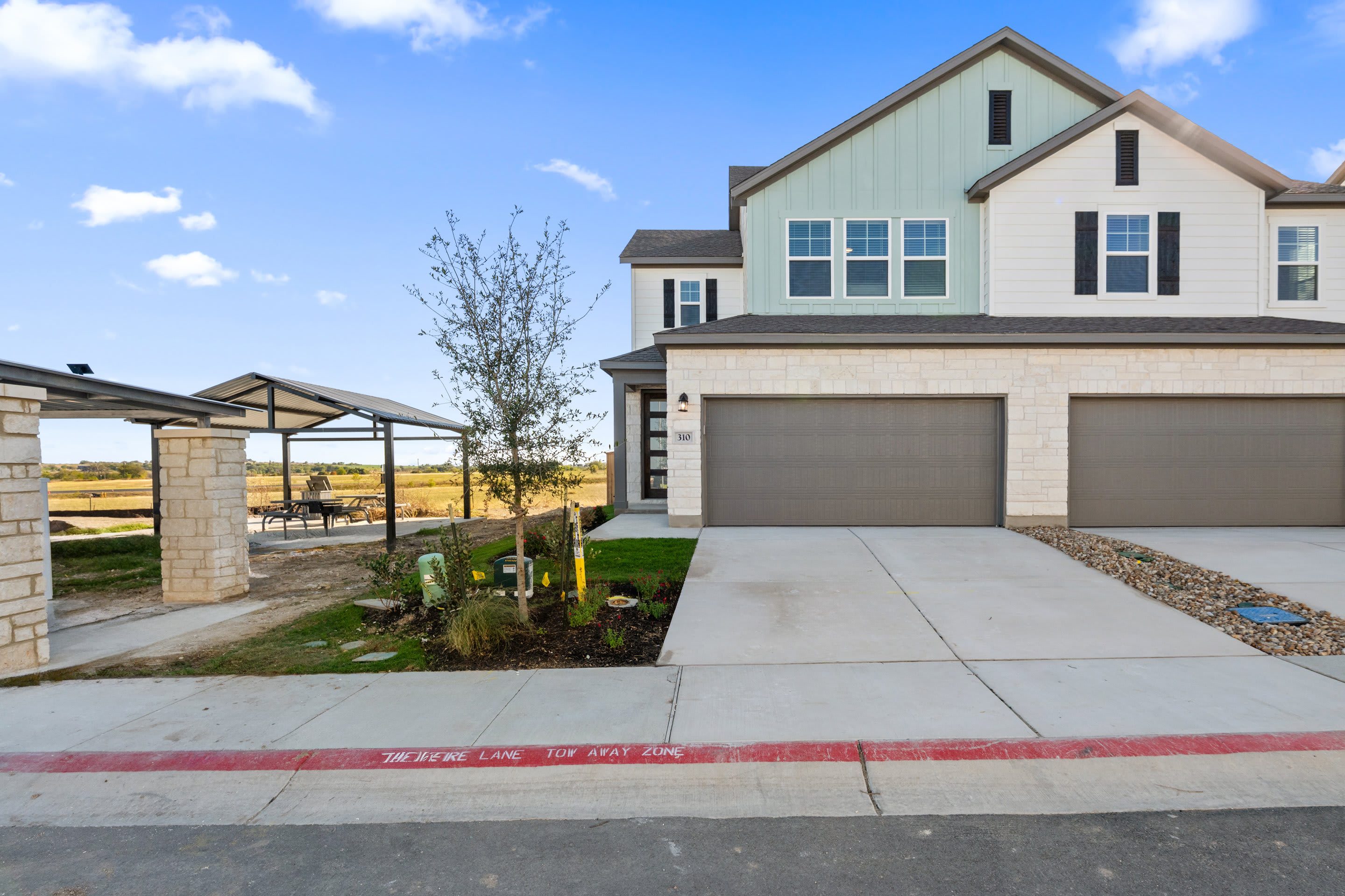 A modern two-story house with a garage, surrounded by a paved driveway, landscaping, and a clear blue sky with scattered clouds in the background.