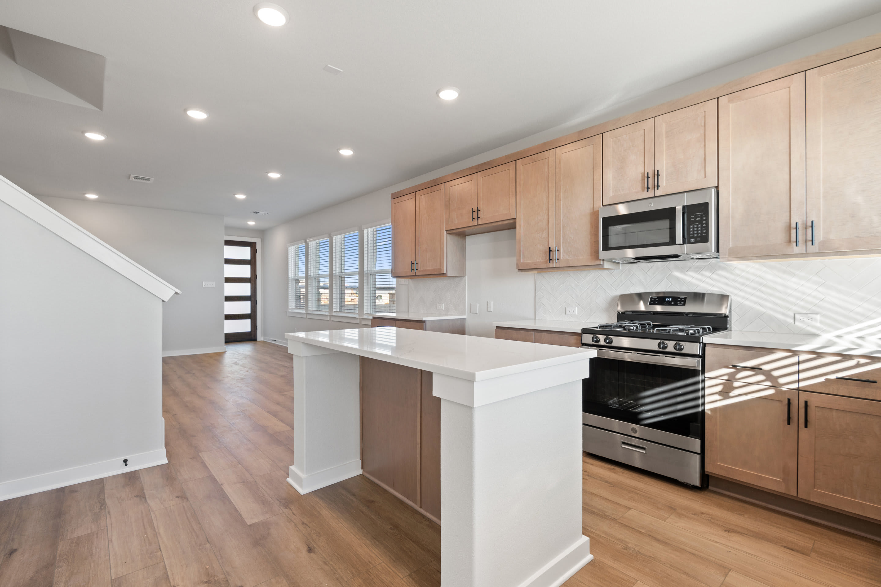 A modern, open-concept kitchen with light wood cabinets, stainless steel appliances, and a white countertop, set against a backdrop of hardwood floors and bright windows.