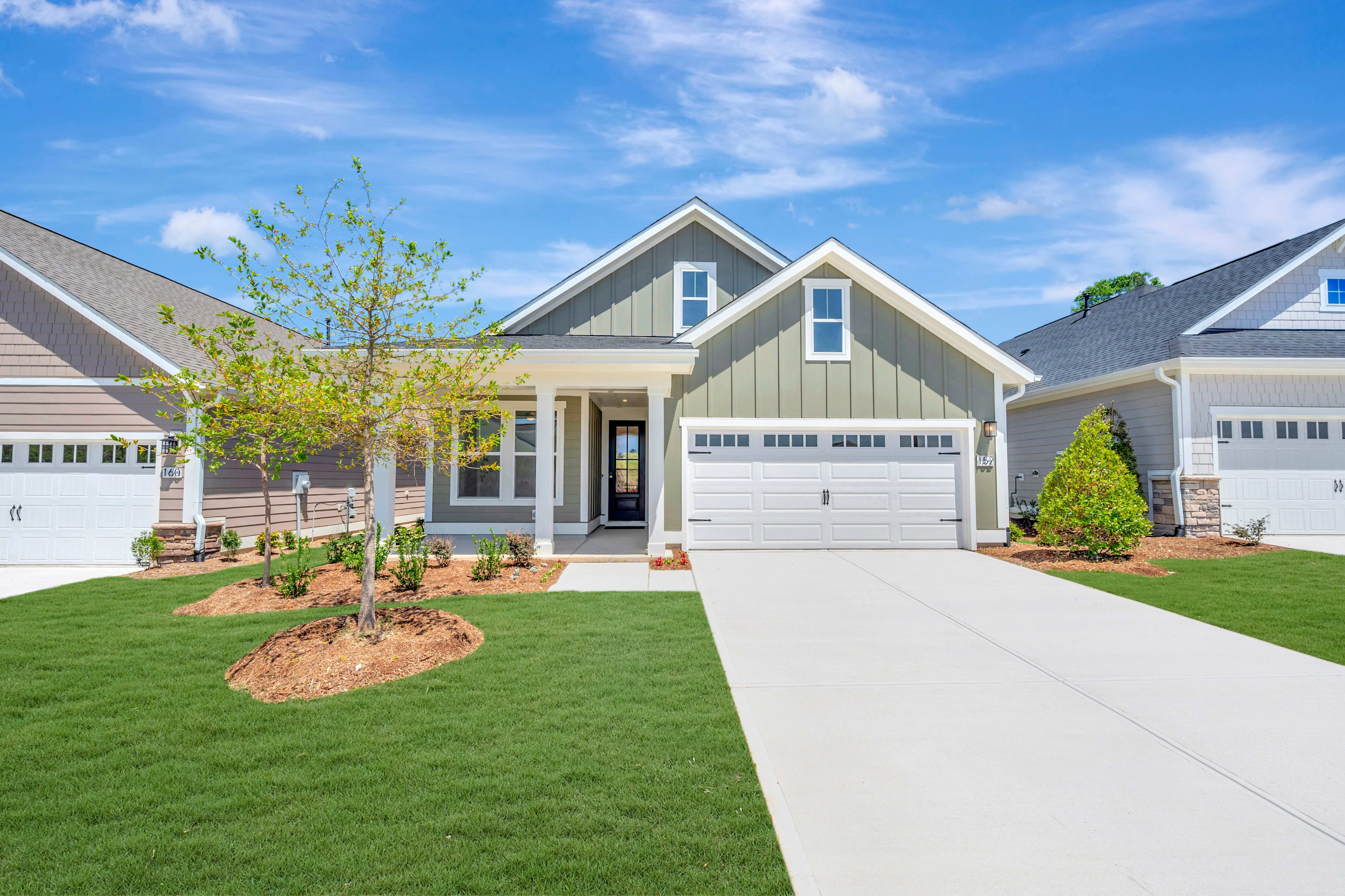 A well-manicured lawn surrounds a modern, two-story house with a white exterior, a covered porch, and a neatly landscaped yard under a bright, blue sky with scattered clouds.