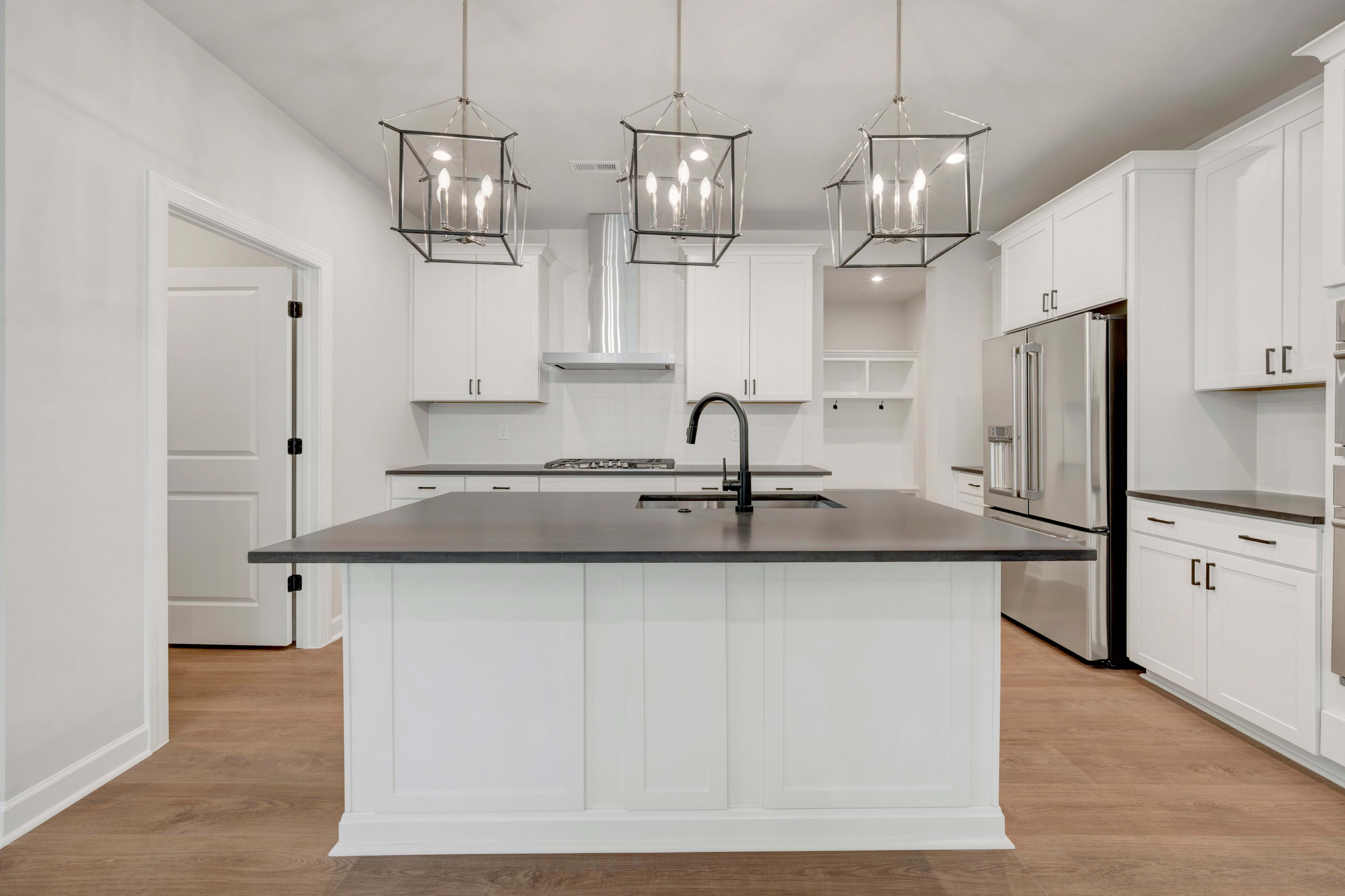 A modern, minimalist kitchen with white cabinets, a large island with a dark countertop, and three pendant lights hanging above.