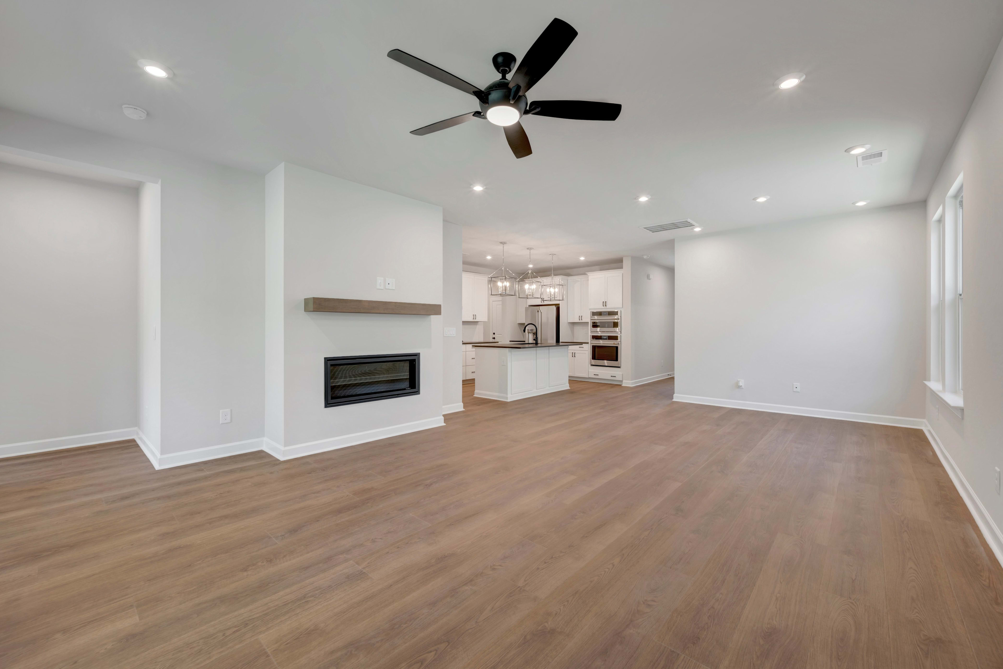 A spacious, well-lit room with a ceiling fan, hardwood floors, and an open kitchen visible in the background.