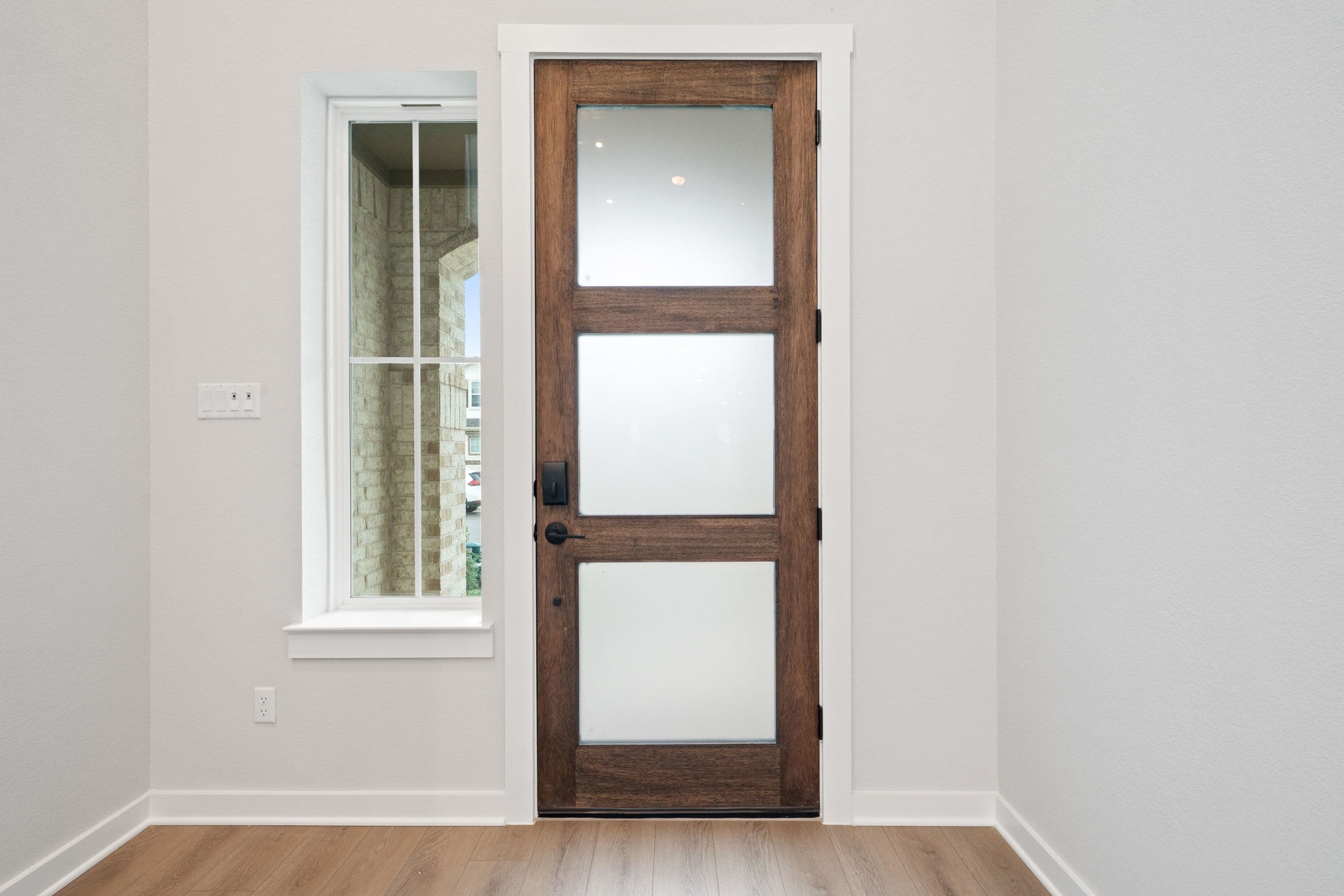 A wooden door with three glass panels stands in a bright, minimalist room with a window visible in the background.