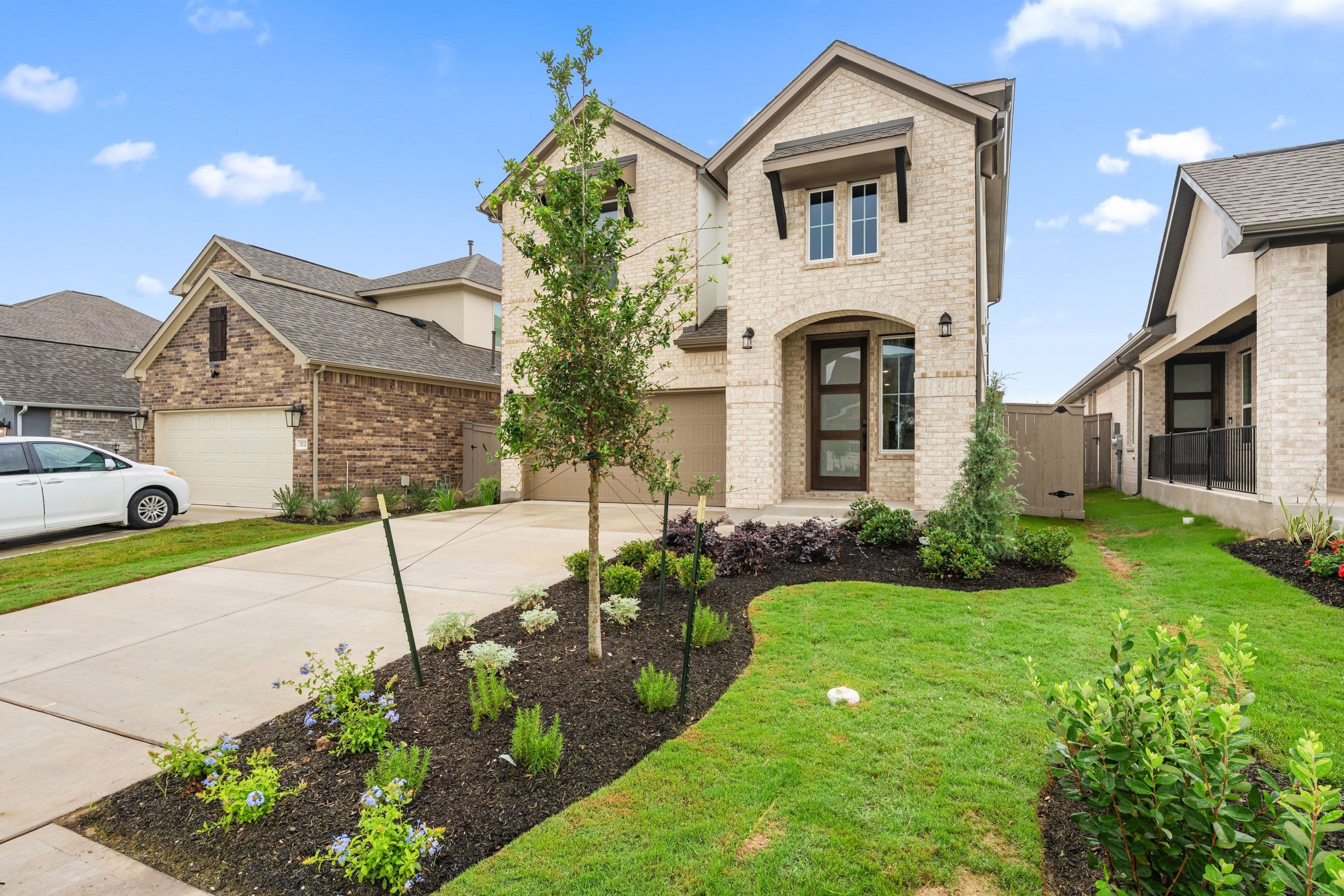 A well-manicured lawn with lush greenery surrounds a stately two-story brick and stone house with a prominent entryway and large windows, set against a clear blue sky with scattered clouds.