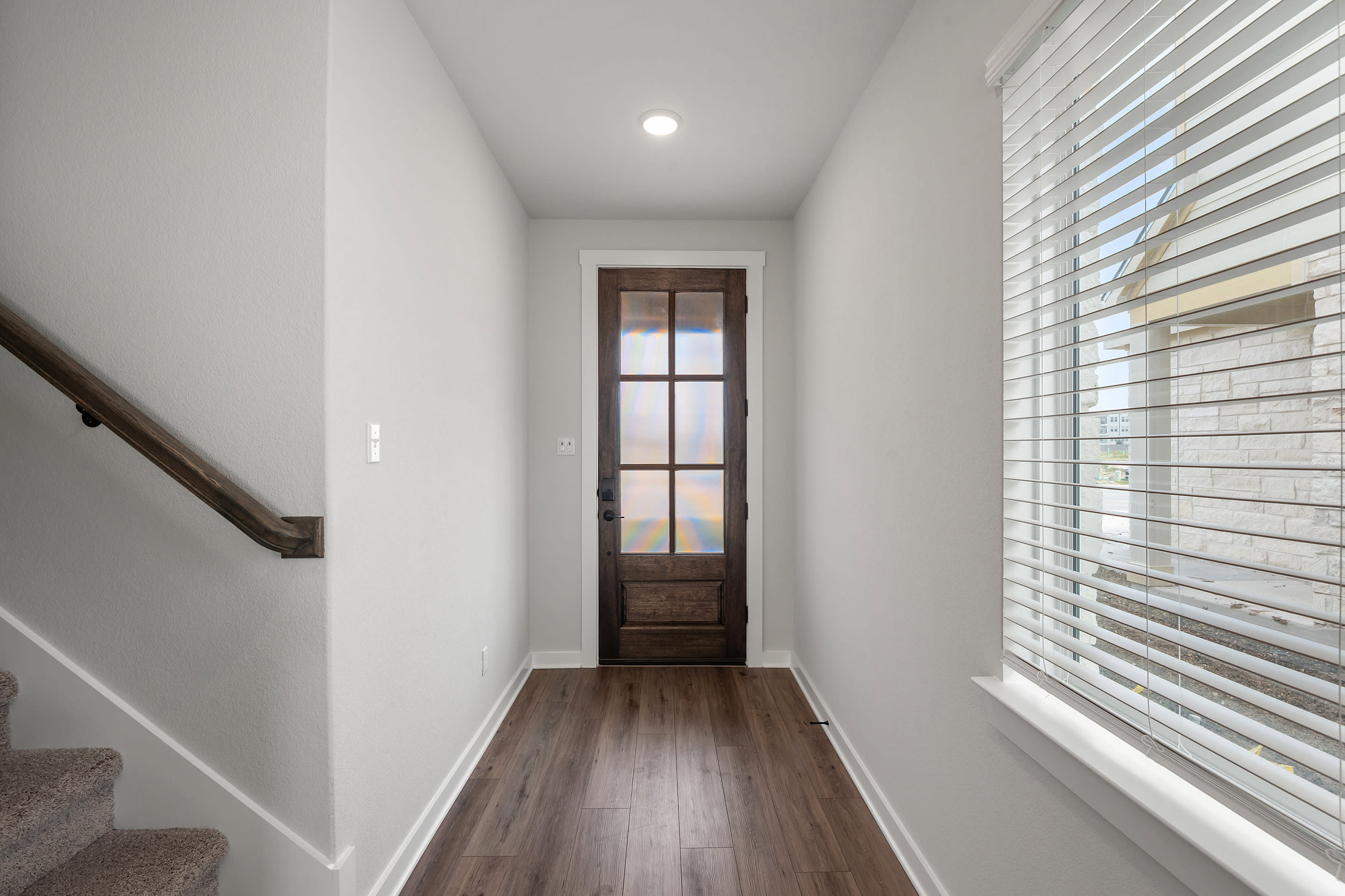 A dark wooden front door is visible at the end of a hallway with a carpeted staircase on the left and a window with blinds on the right.