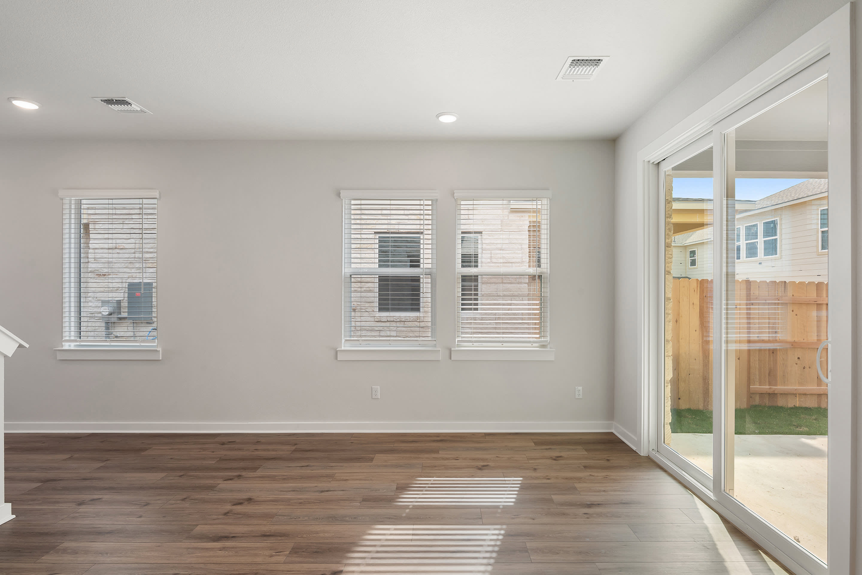 A bright, empty living room with wood-look flooring in the foreground opens to a backyard with a wooden fence and houses in the background.