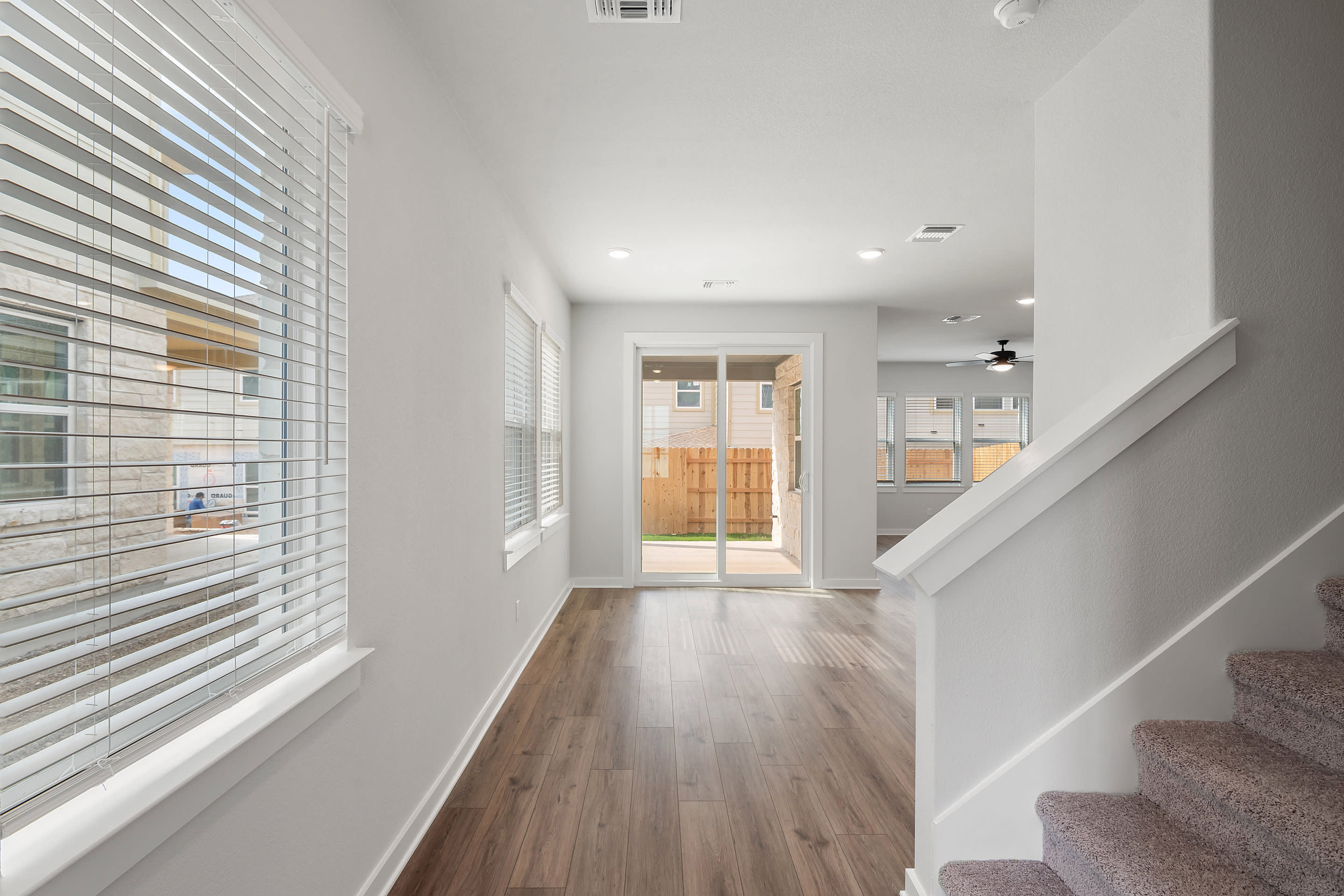 A bright, modern living room with a wooden floor and a staircase in the foreground opens to a backyard visible through sliding glass doors in the background.