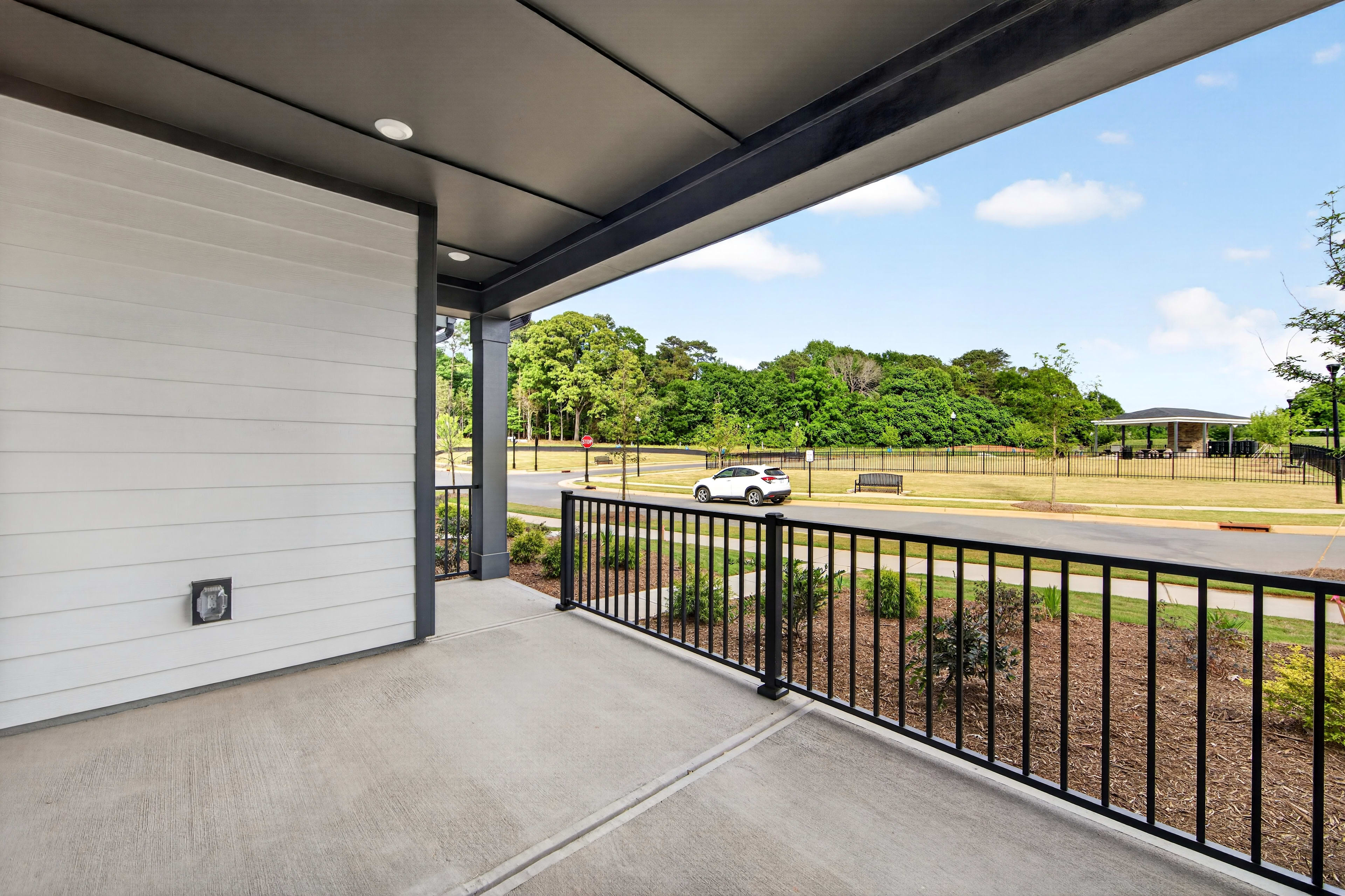 A modern porch with a white wall and black railing overlooks a grassy park with trees and a white car.