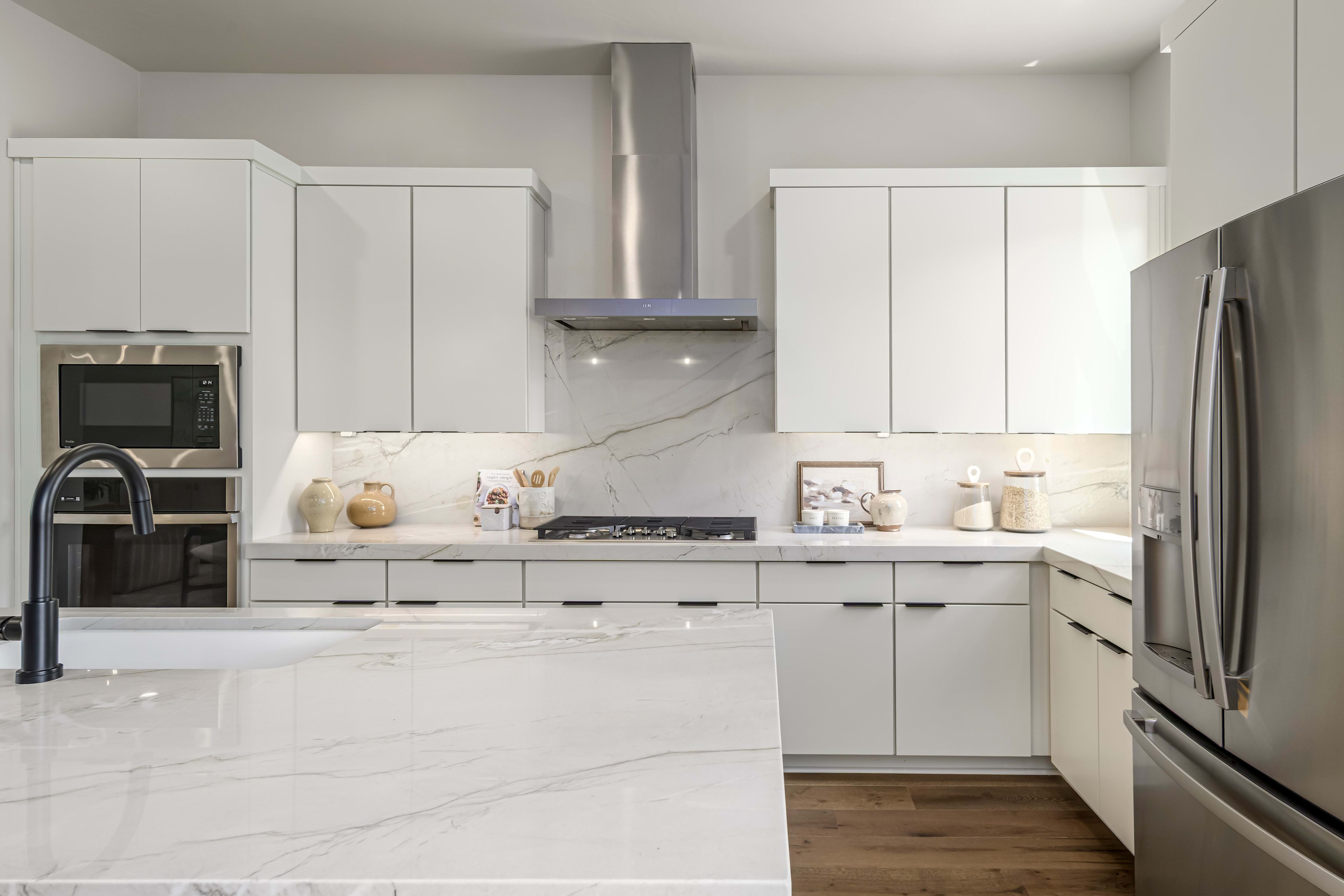 A modern white kitchen with a marble island in the foreground and stainless steel appliances and cabinetry in the background.