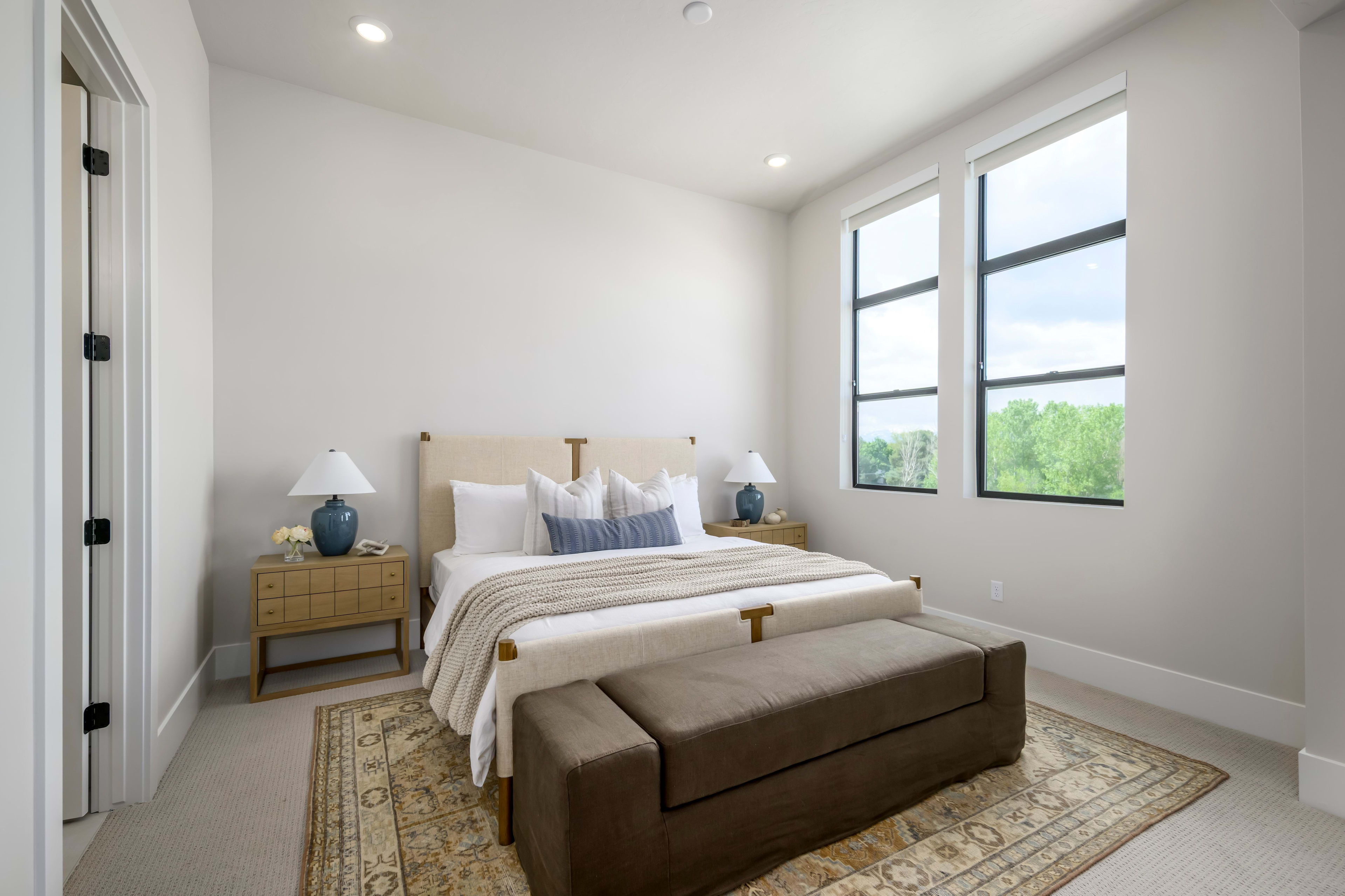 A neatly made bed with a bench at its foot sits on an ornate rug in the foreground, while large windows in the background reveal a lush green landscape.