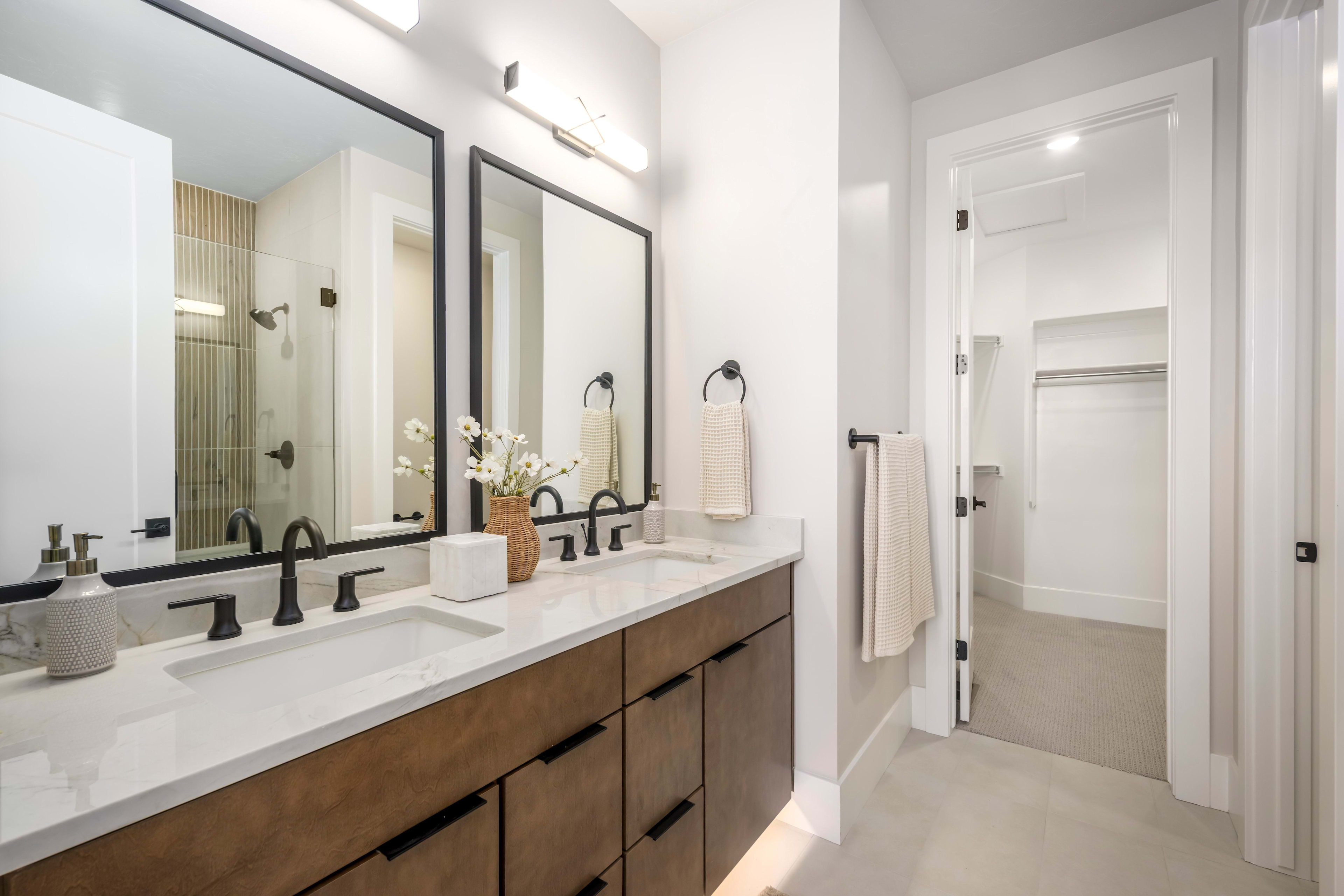 A modern double vanity with black fixtures and a wood-grain cabinet sits in the foreground, while a walk-in closet is visible through an open doorway in the background.