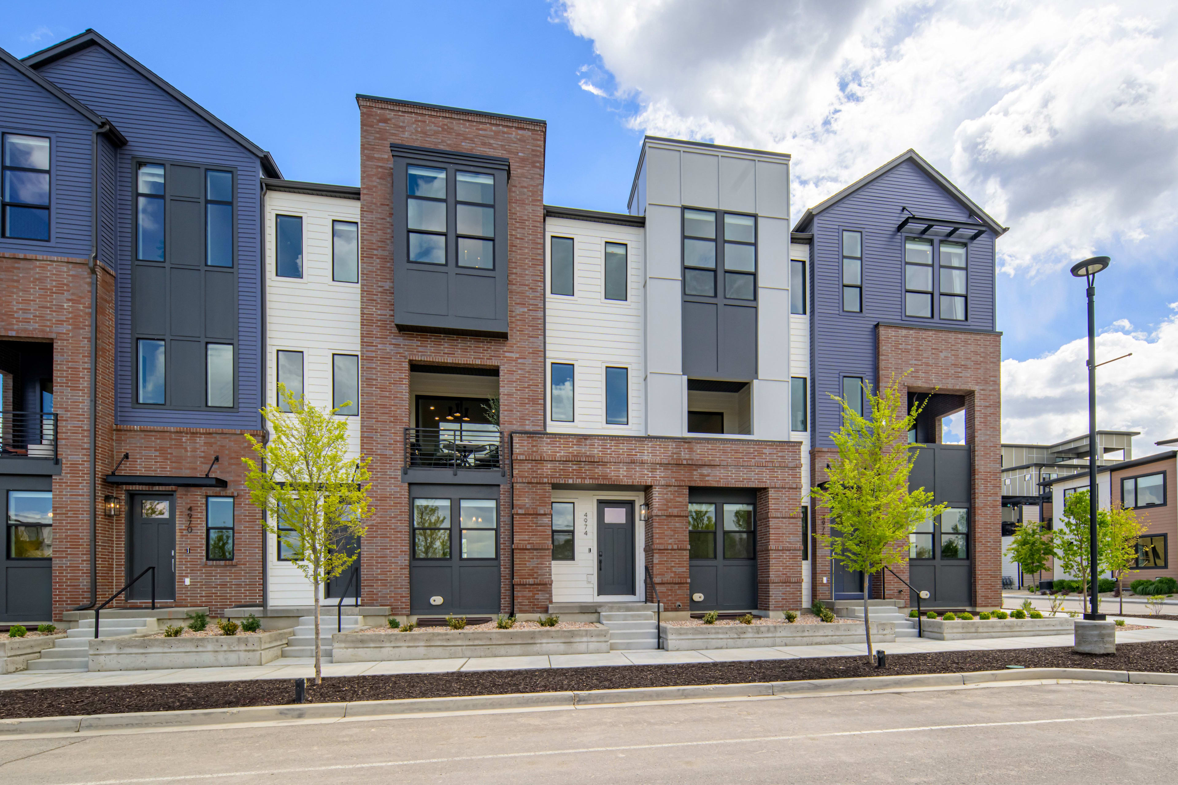 Modern townhouses with brick and white siding stand in the foreground, while a street and more buildings are visible in the background under a cloudy sky.