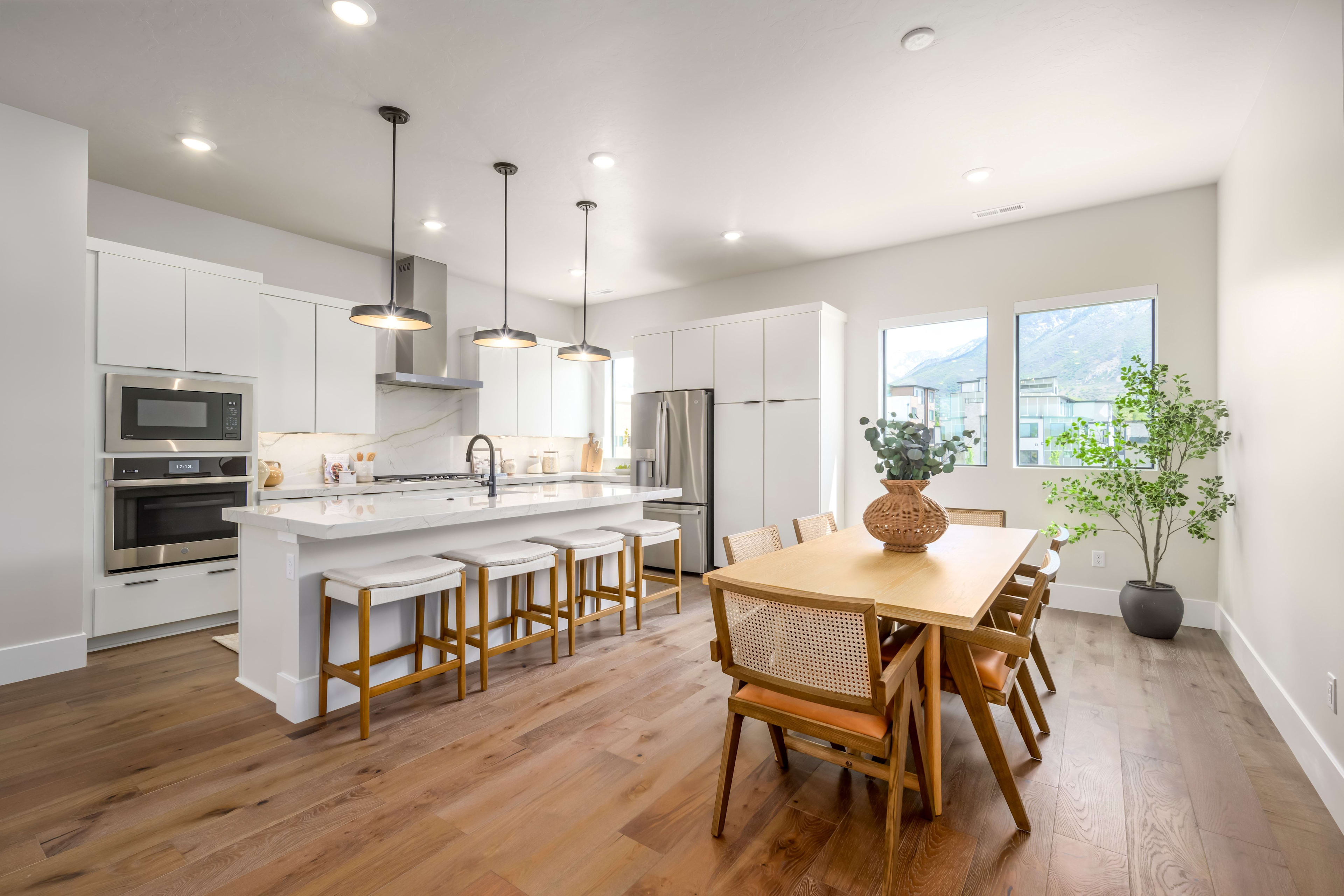 A modern white kitchen with a large island and bar stools is in the foreground, while a dining table with chairs and a potted plant are in the background, with windows revealing a view of houses and hills.