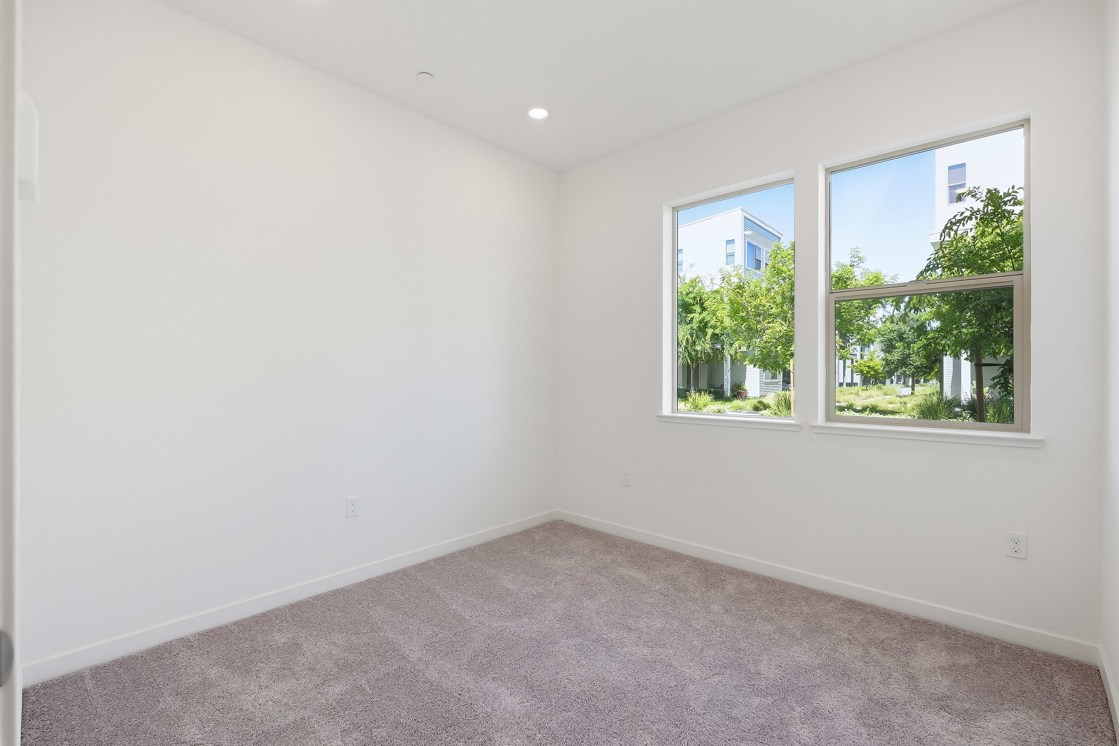 A carpeted room with white walls and two windows looks out onto a sunny street lined with trees and modern buildings.