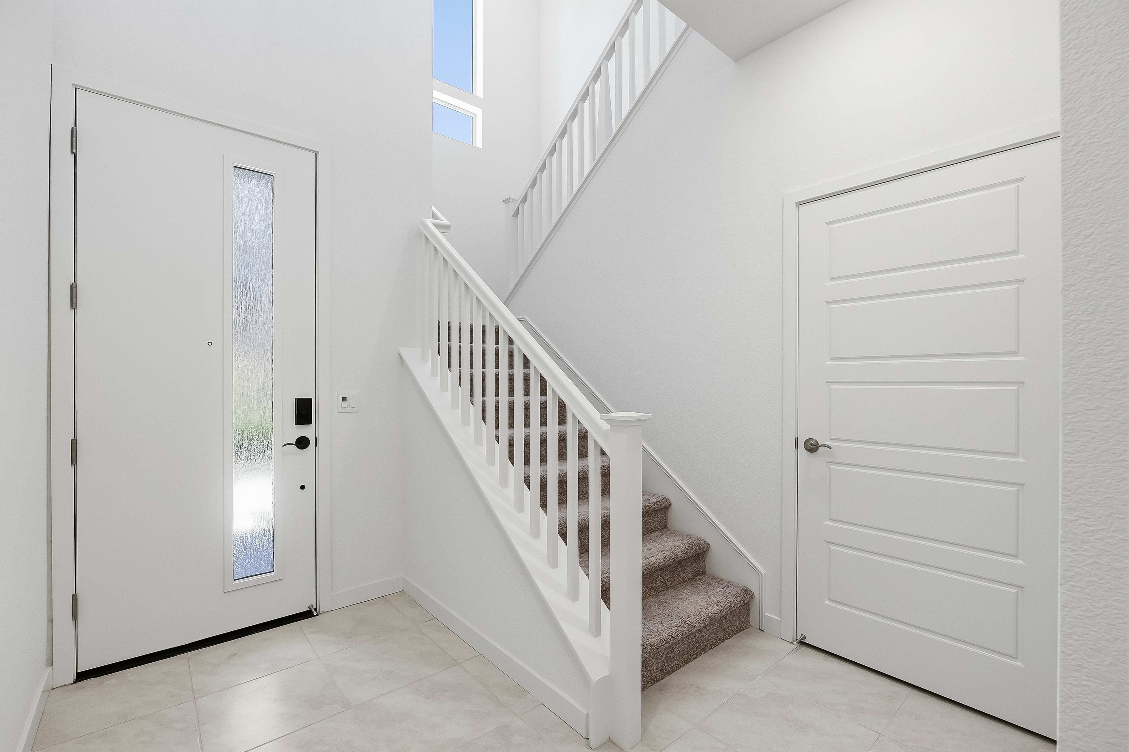 A bright white entryway features a modern staircase with carpeted steps in the foreground, leading up to a large window and a white door on the right in the background.