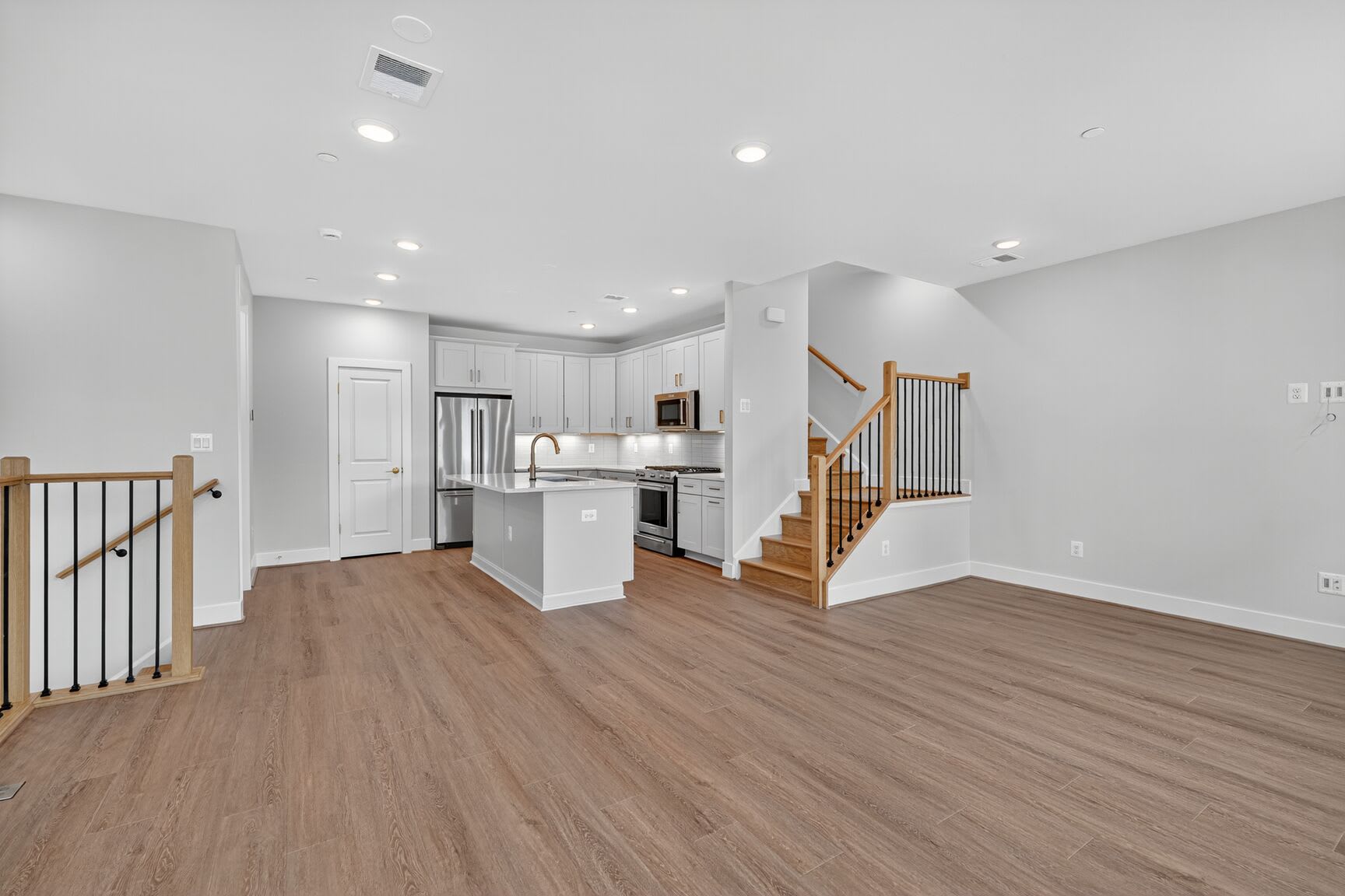 A modern kitchen with a central island and stainless steel appliances is visible in the background, while the foreground features a spacious living area with hardwood floors and a staircase.