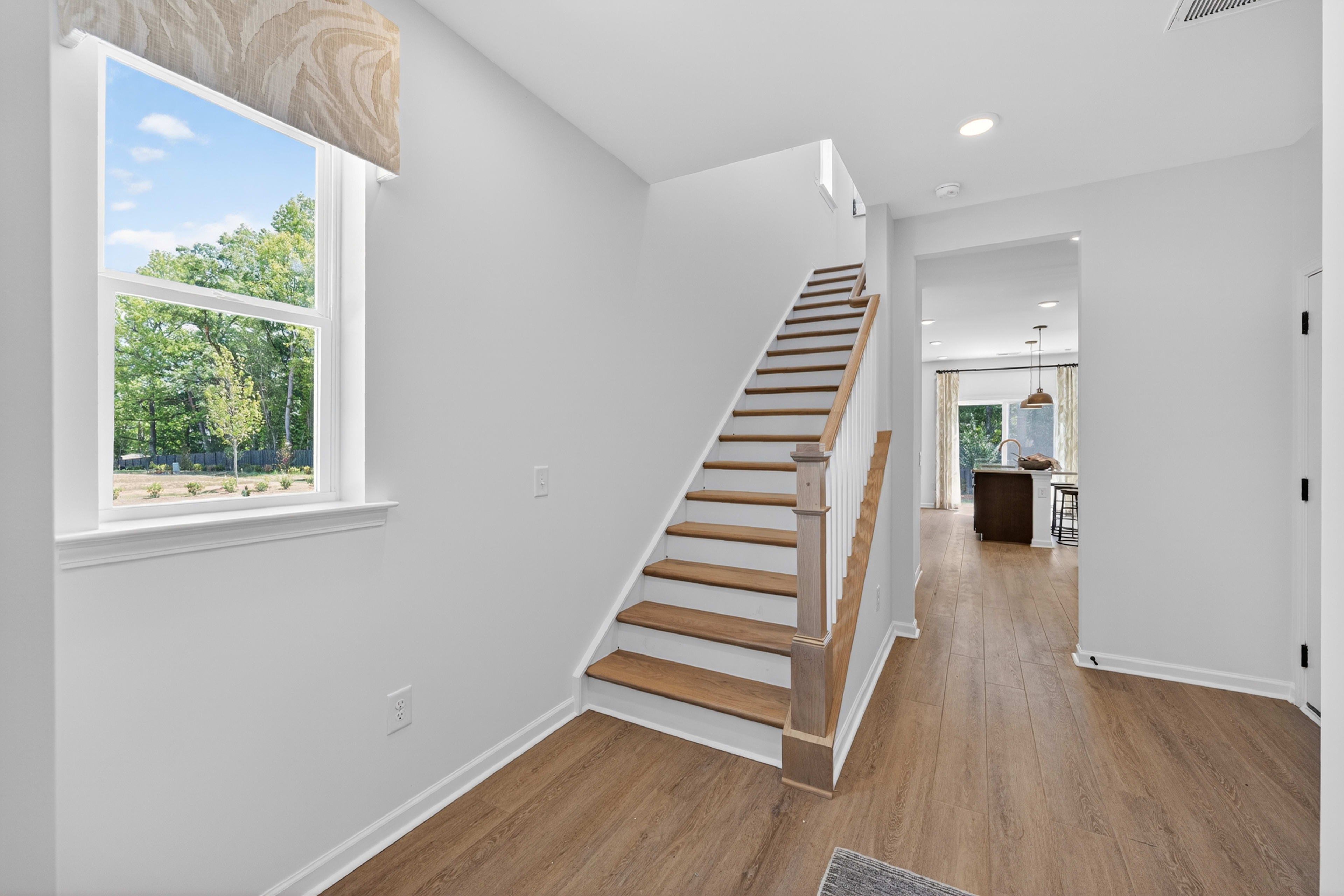 A bright staircase with wooden steps and railing leads to an open living area with a kitchen island, while a large window in the foreground offers a view of green trees under a blue sky.
