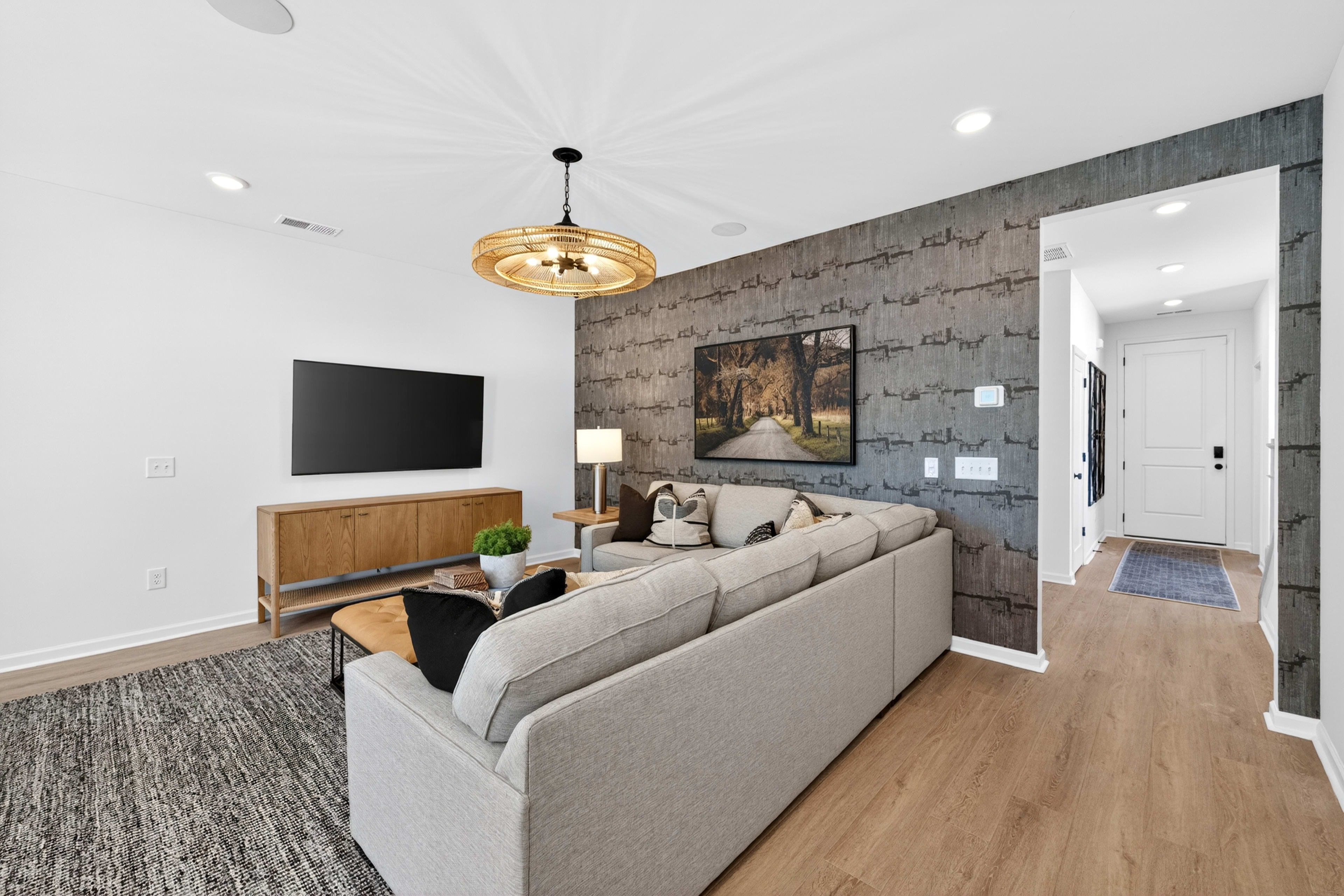 A modern living room with a sectional sofa and a television in the foreground, and a hallway leading to a front door in the background.