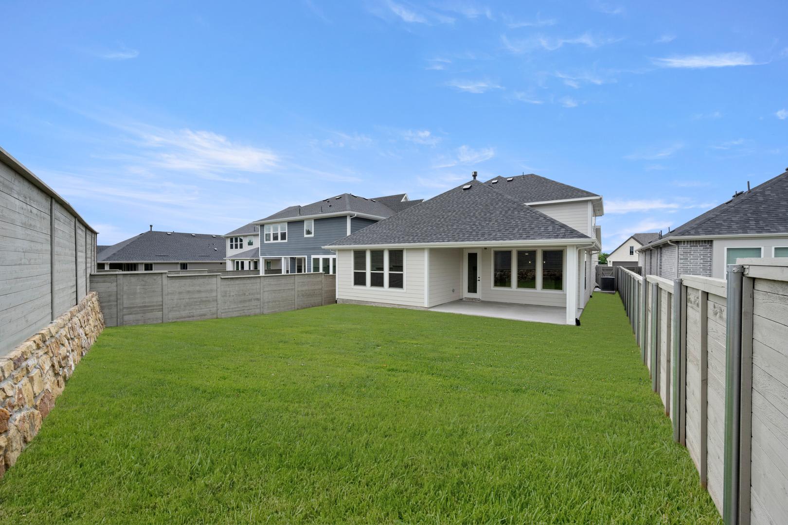 A lush green lawn fills the foreground, bordered by a stone and concrete fence, while modern houses with dark roofs stand in the background under a clear blue sky.