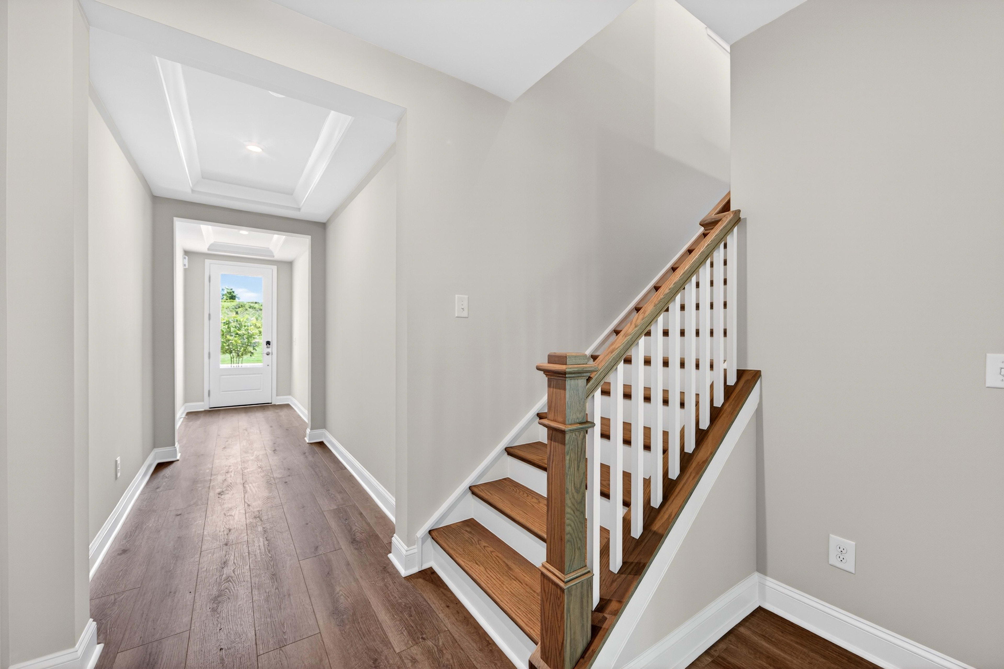 A modern hallway with a wooden floor leads to a bright doorway in the background, while a staircase with white spindles and wooden steps occupies the right side of the foreground.