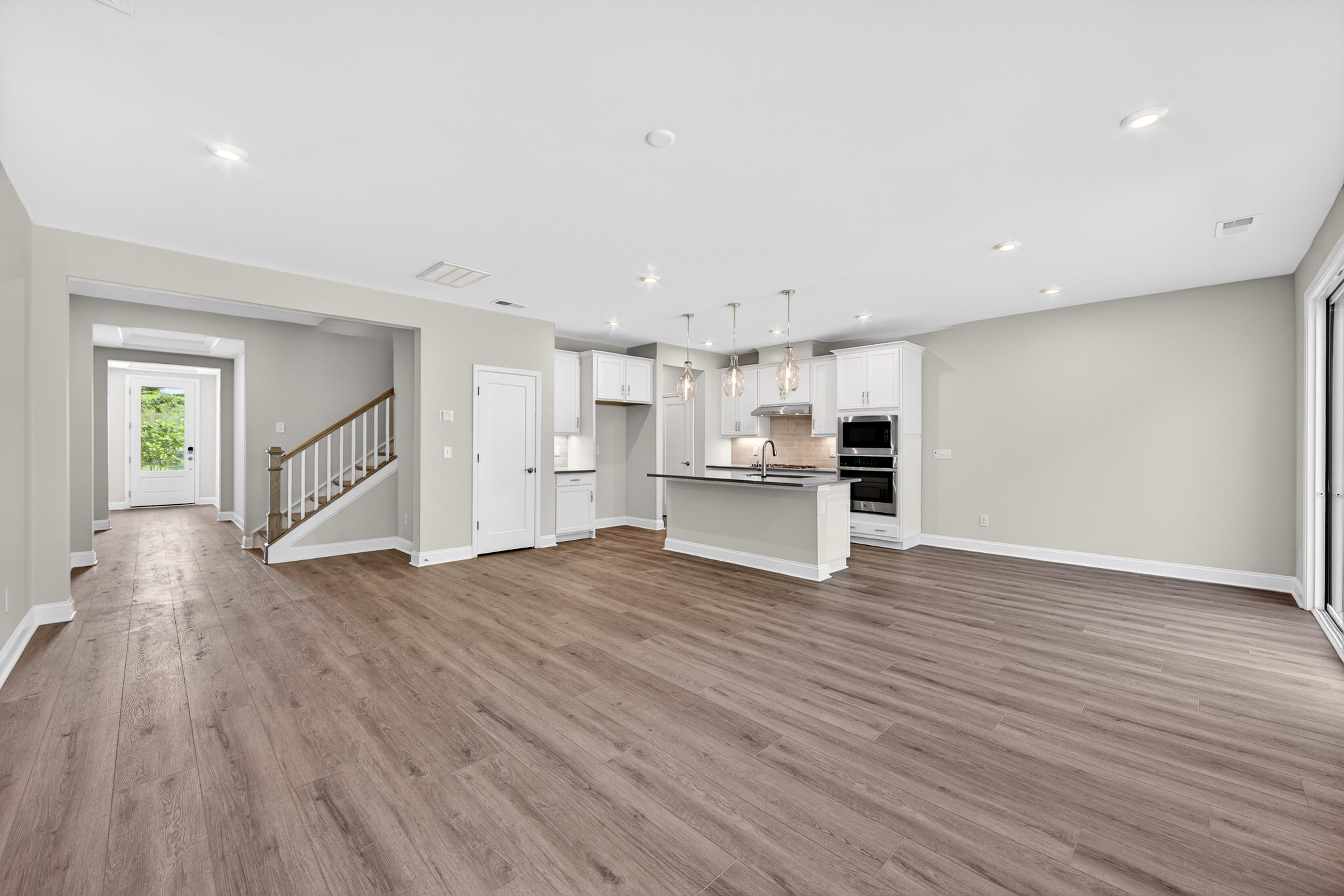 A spacious, modern open-plan living area with a light wood floor in the foreground leads to a white kitchen with an island and stainless steel appliances in the background, with a staircase and doorway visible to the left.