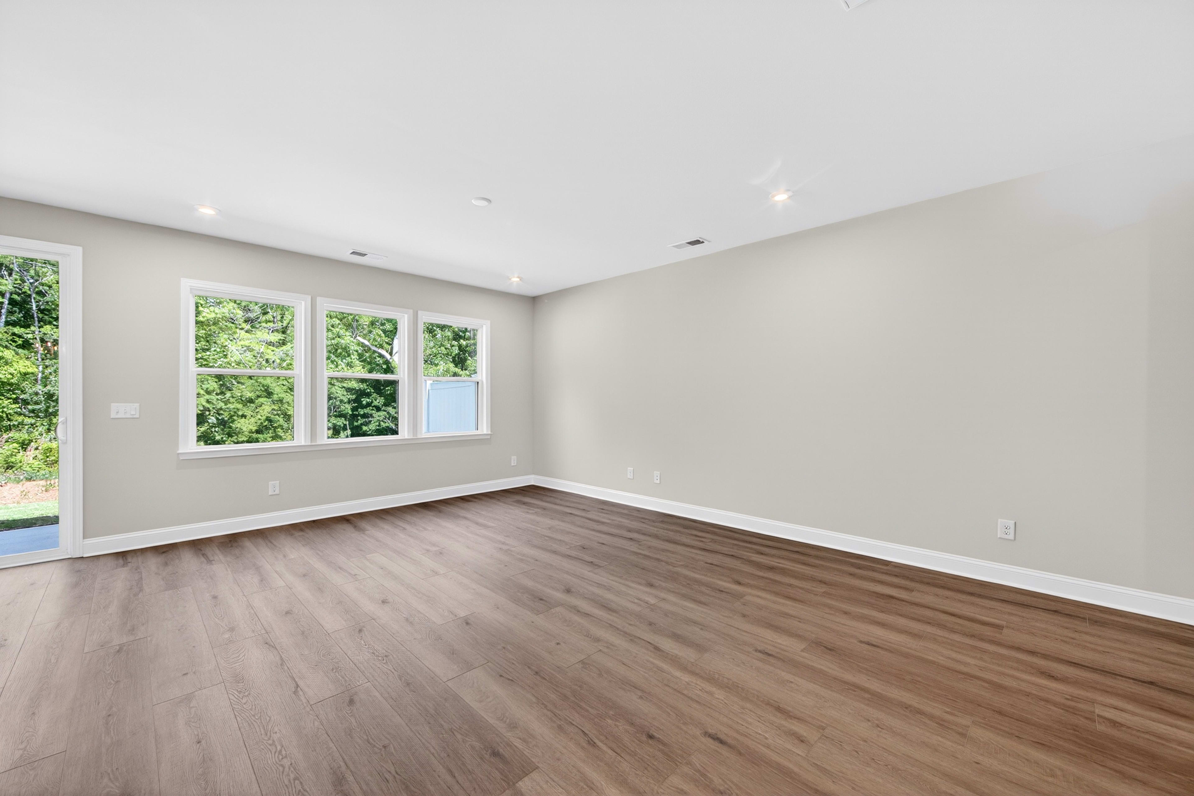 A spacious, empty room with dark wood flooring in the foreground and large windows overlooking lush green trees in the background.