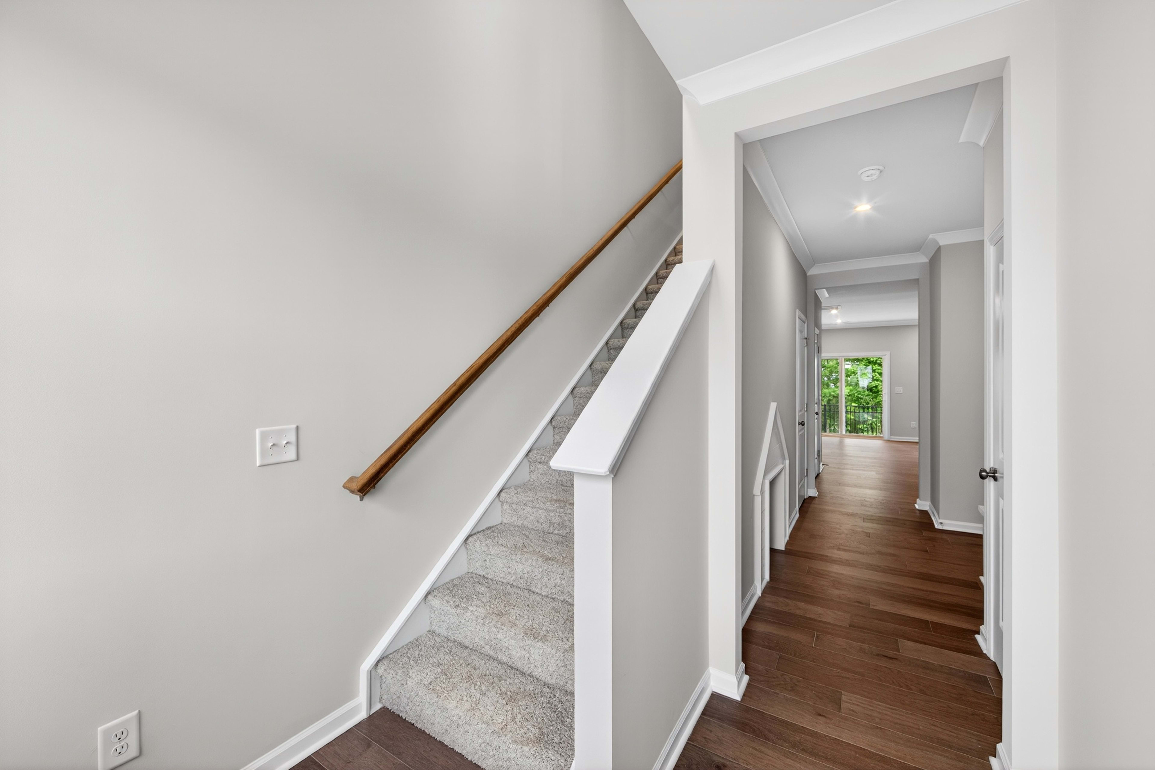 A carpeted staircase with a wooden handrail is in the foreground, leading to a hallway with dark wood floors and doors in the background.