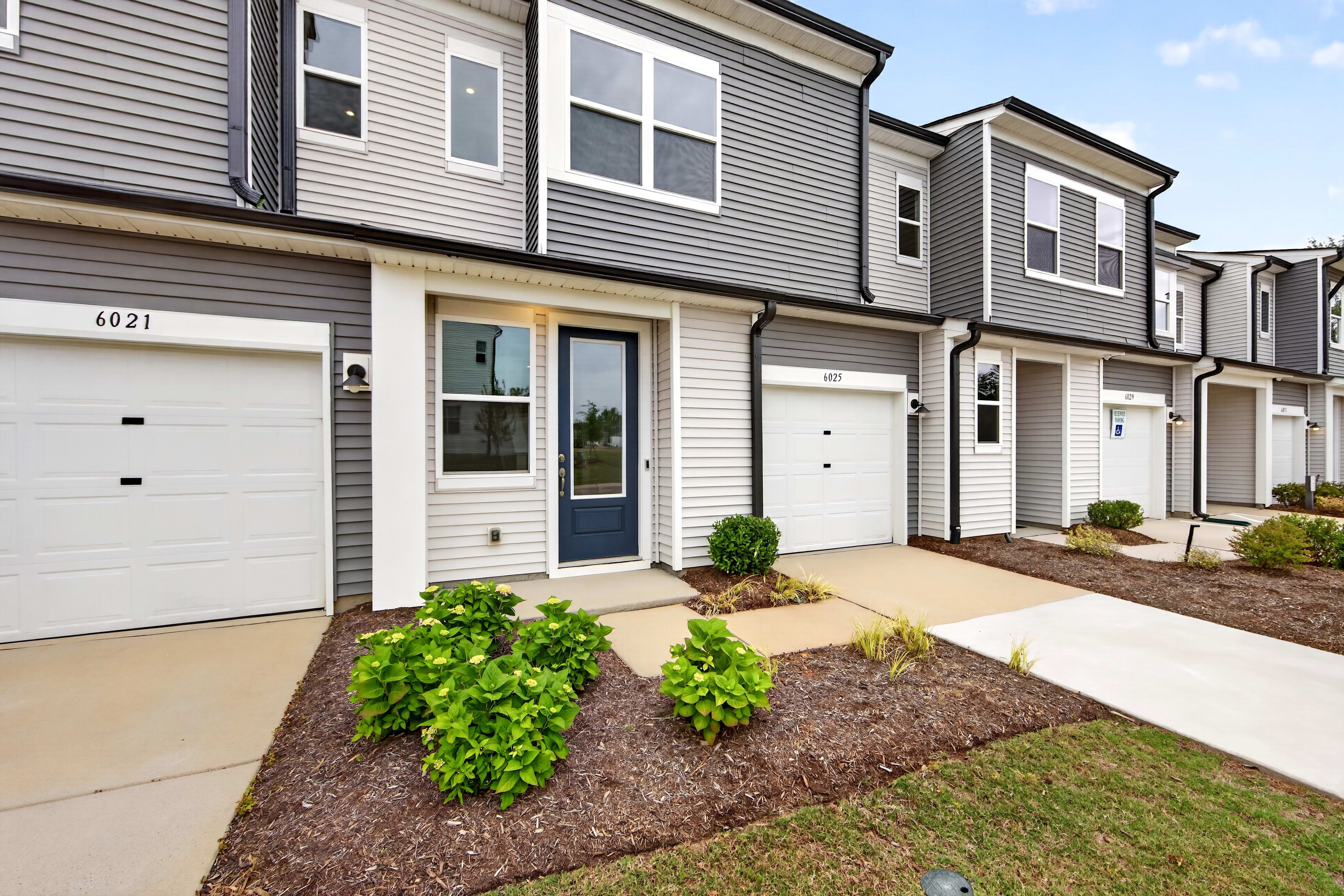 Lush green bushes and mulch fill the foreground, leading to a row of modern townhouses with garages and dark blue doors in the background.