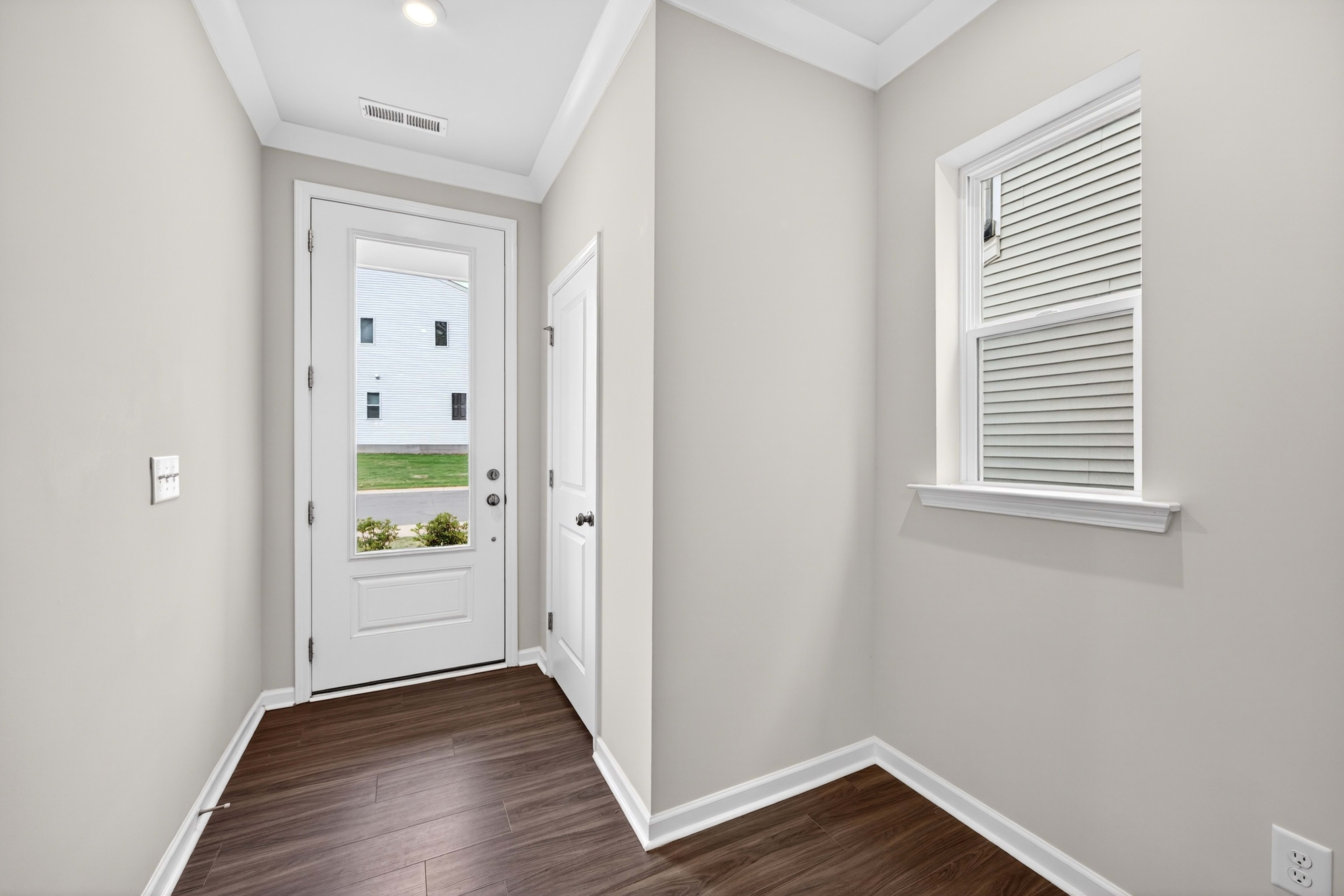 A dark wood floor leads to a glass-paned door with a view of a white building and green grass, while a window with blinds is visible on the right wall.
