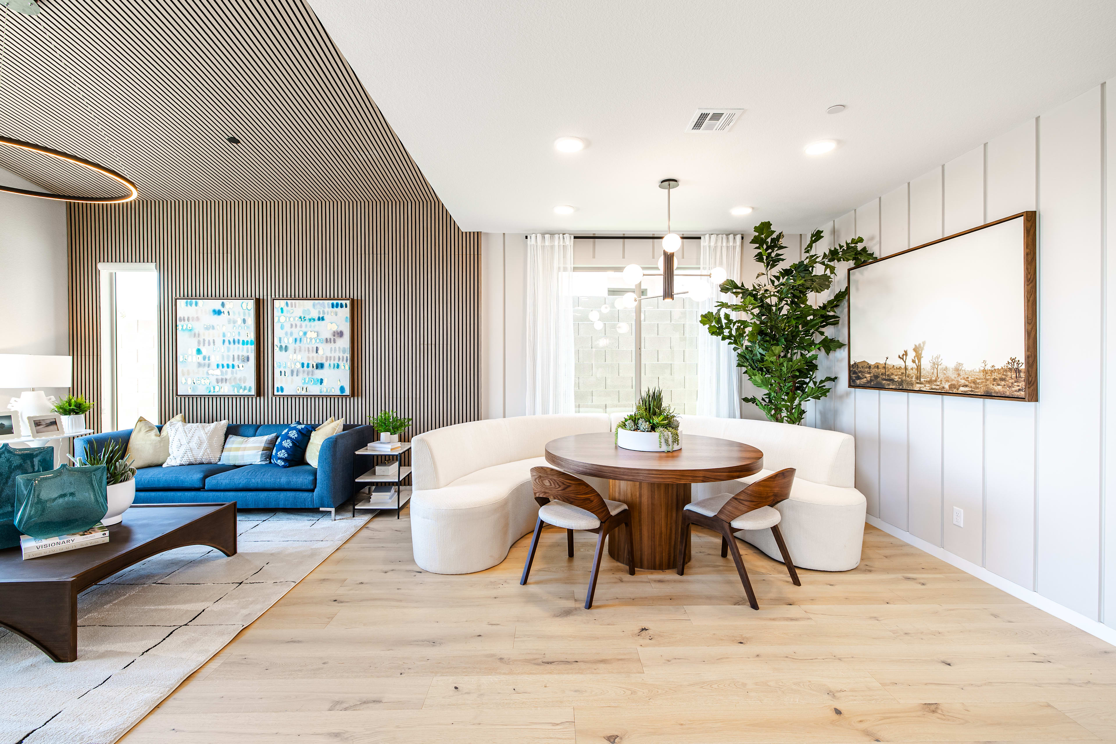 A modern living and dining area features a curved banquette and round table in the foreground, with a blue sofa and artwork in the background.