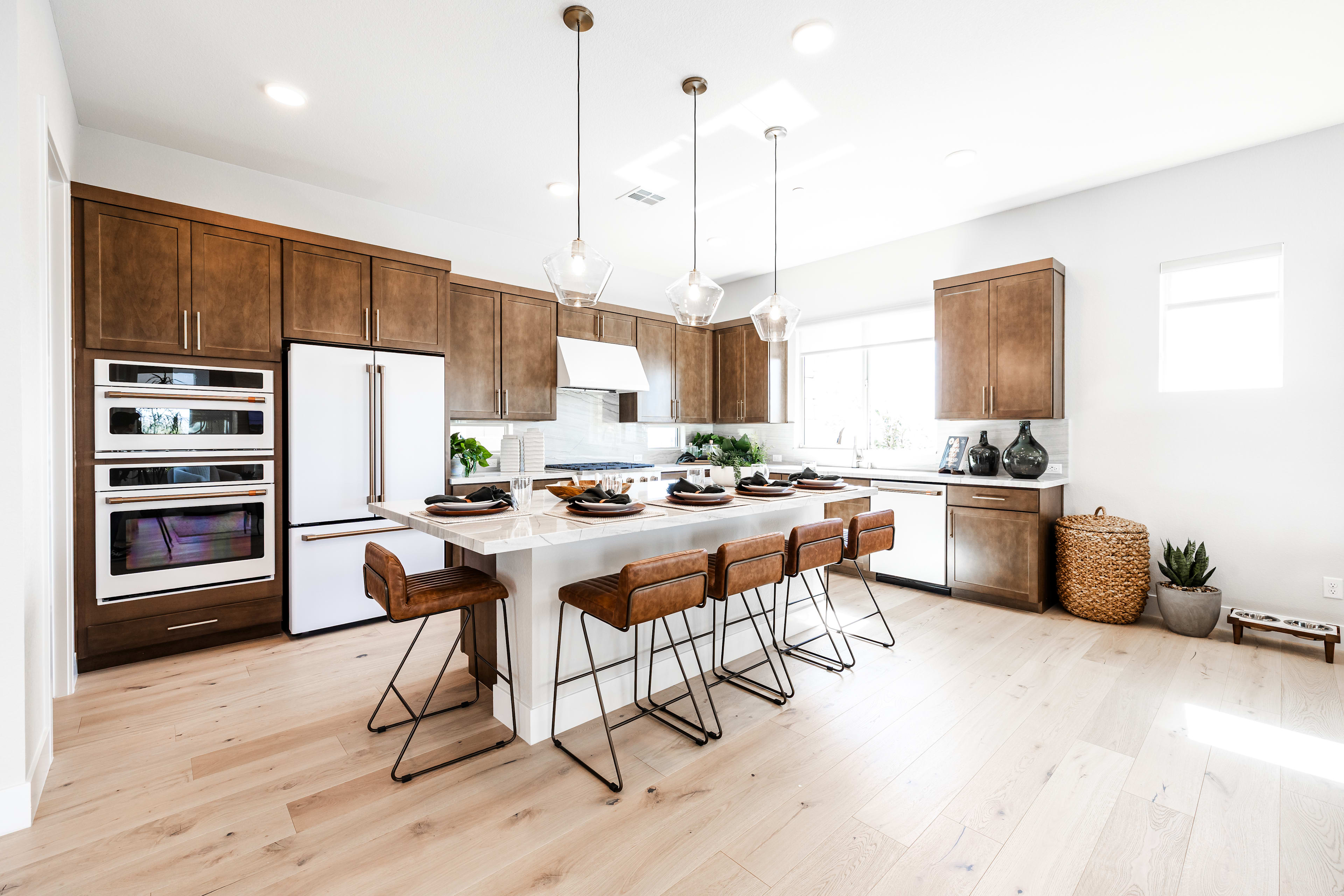 A modern kitchen with a large island and bar stools in the foreground is set against a backdrop of white walls and ample natural light.