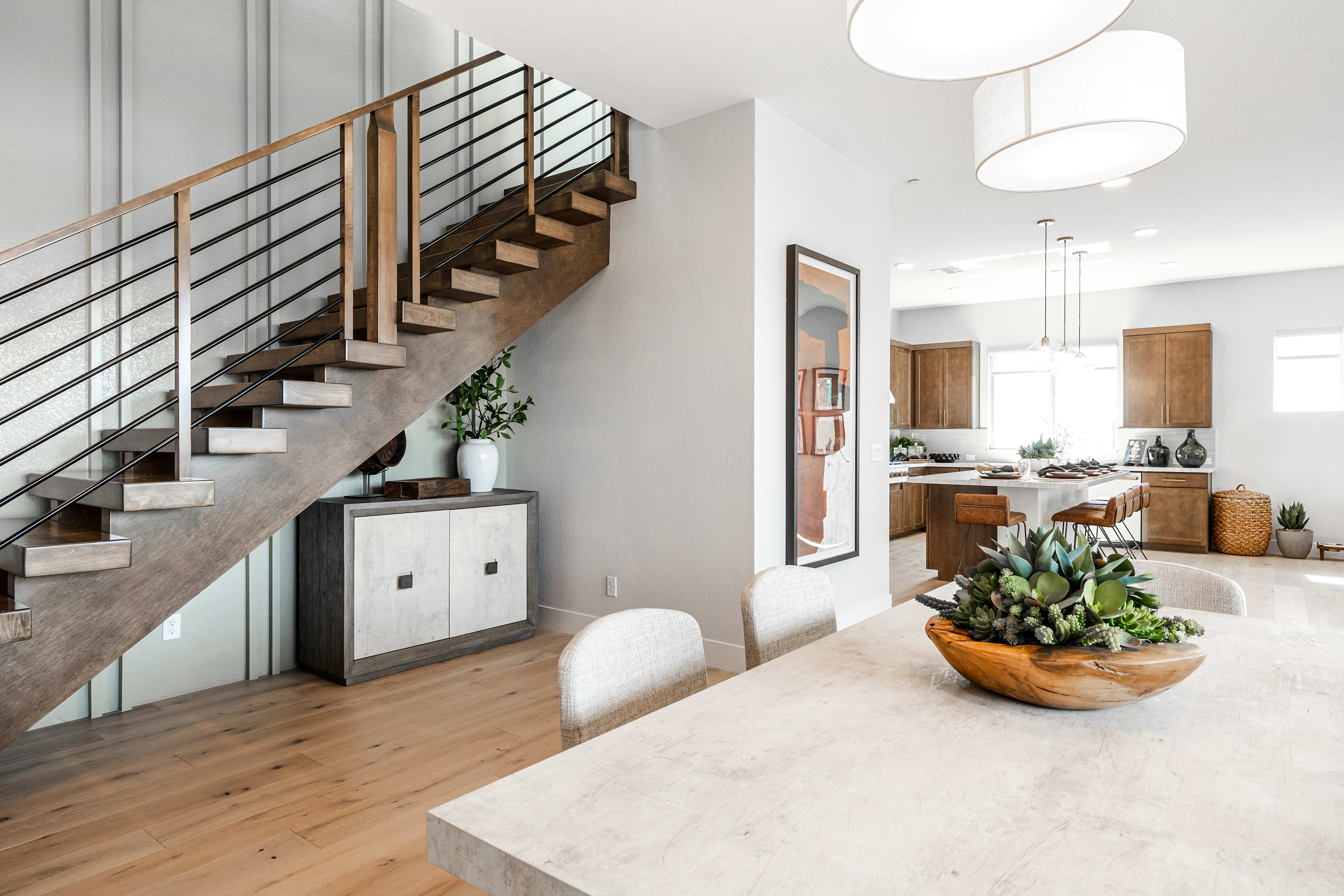 A modern dining area with a concrete table and succulent centerpiece is in the foreground, while a sleek staircase and a glimpse of a kitchen are in the background.