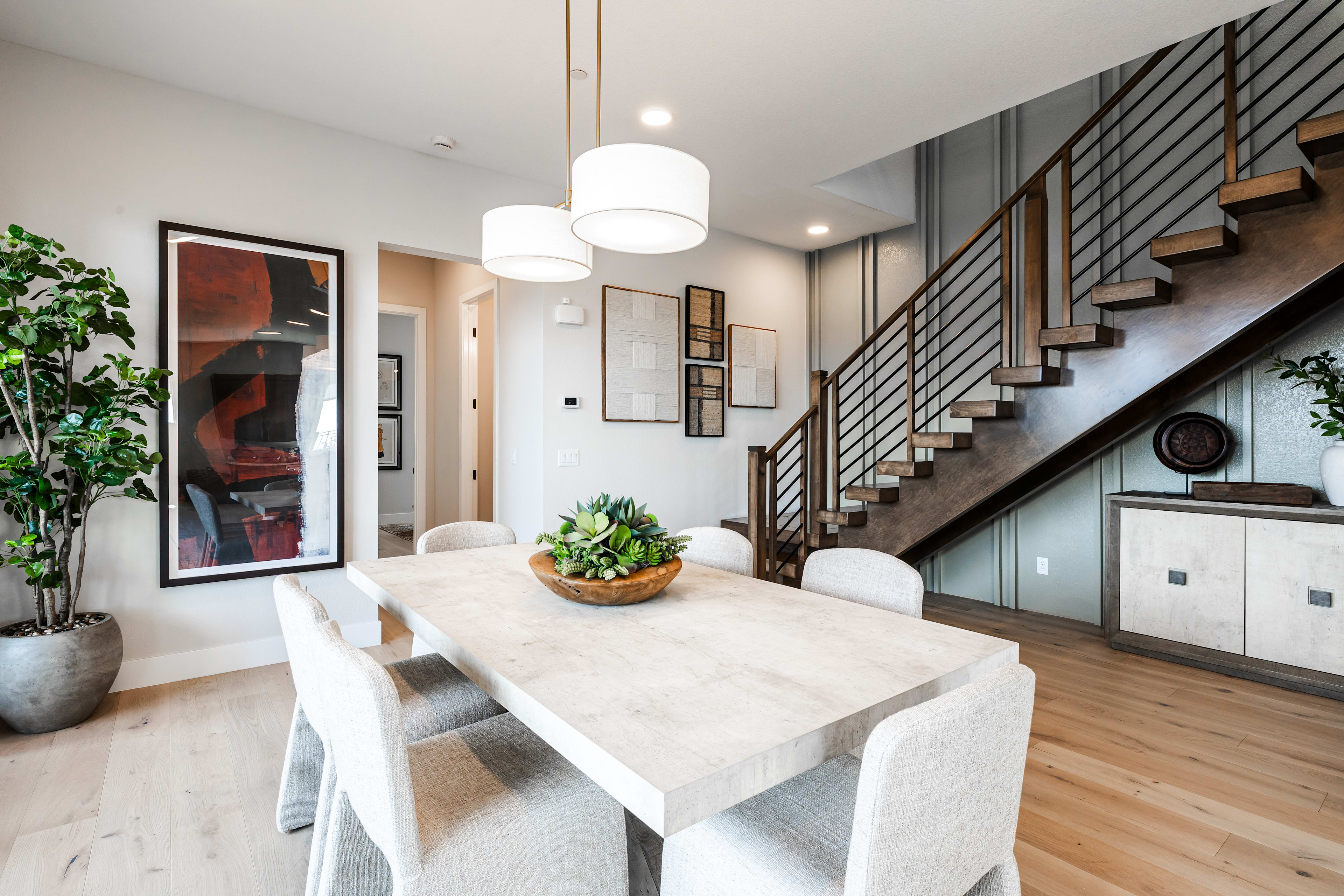 A modern dining room with a light wood table and chairs in the foreground is set against a background featuring a staircase with metal railings and contemporary artwork.