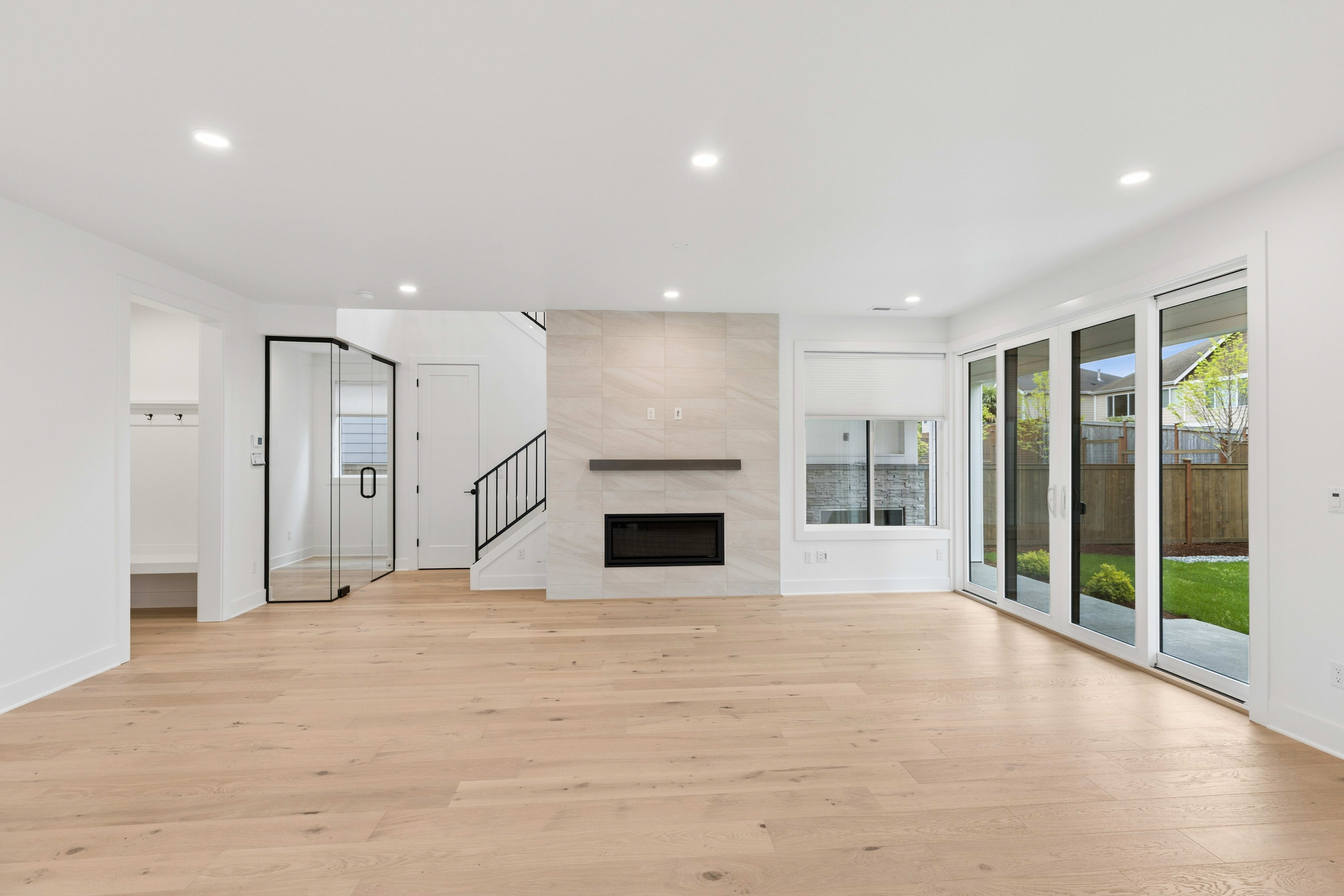 A modern living room with light wood floors in the foreground and a fireplace with a marble surround and large glass doors opening to a green backyard in the background.