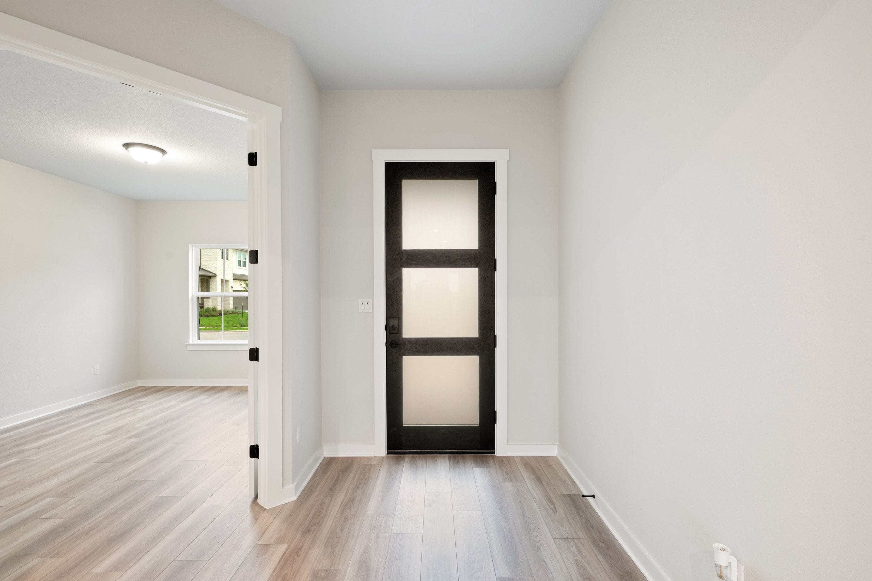 A modern black door with frosted glass panels stands in the background, while the foreground features a light-colored wooden floor and an open doorway leading to a bright room.
