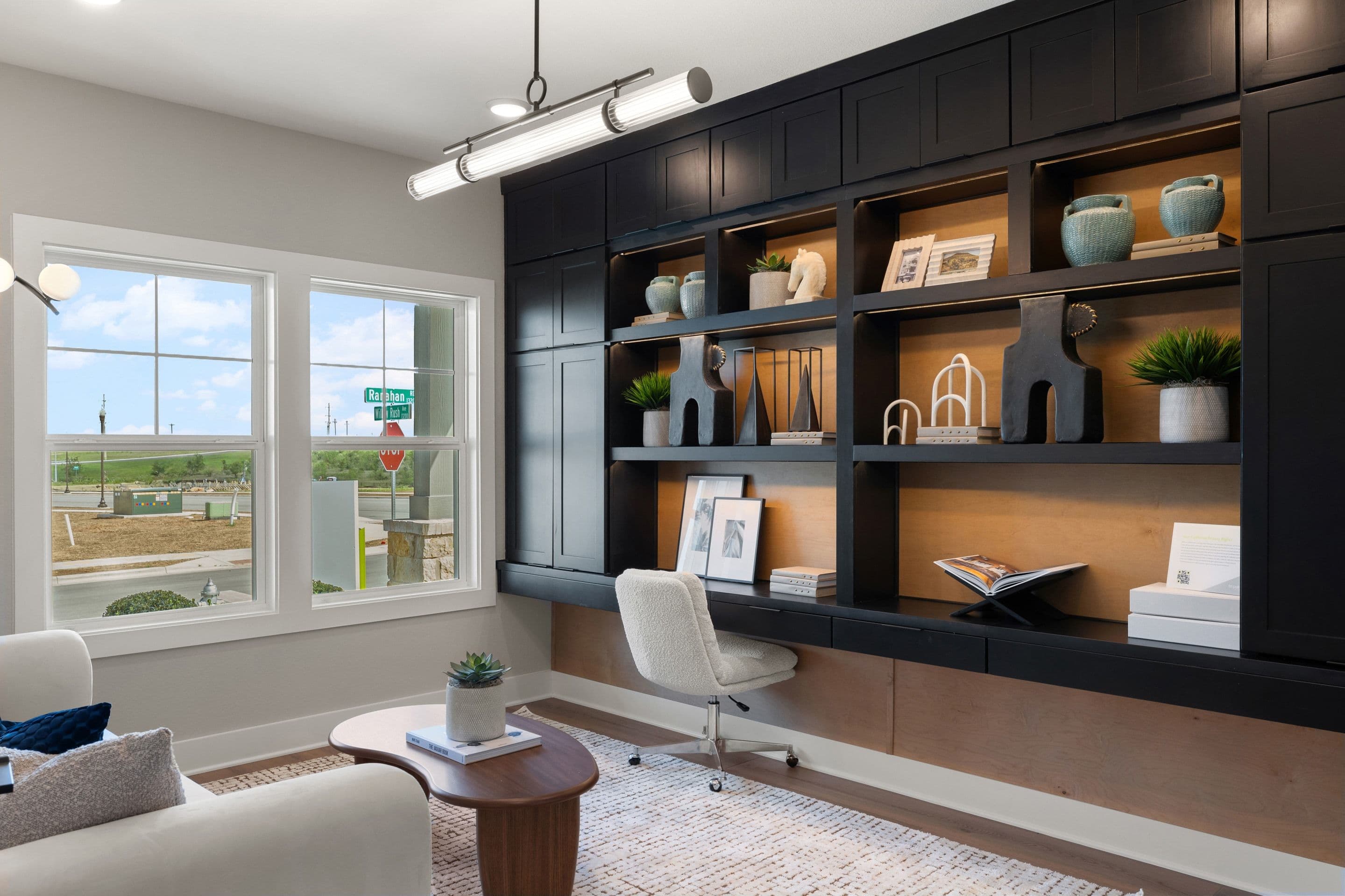 A modern home office with a white desk and chair in front of a dark built-in bookshelf, and large windows overlooking a suburban street.