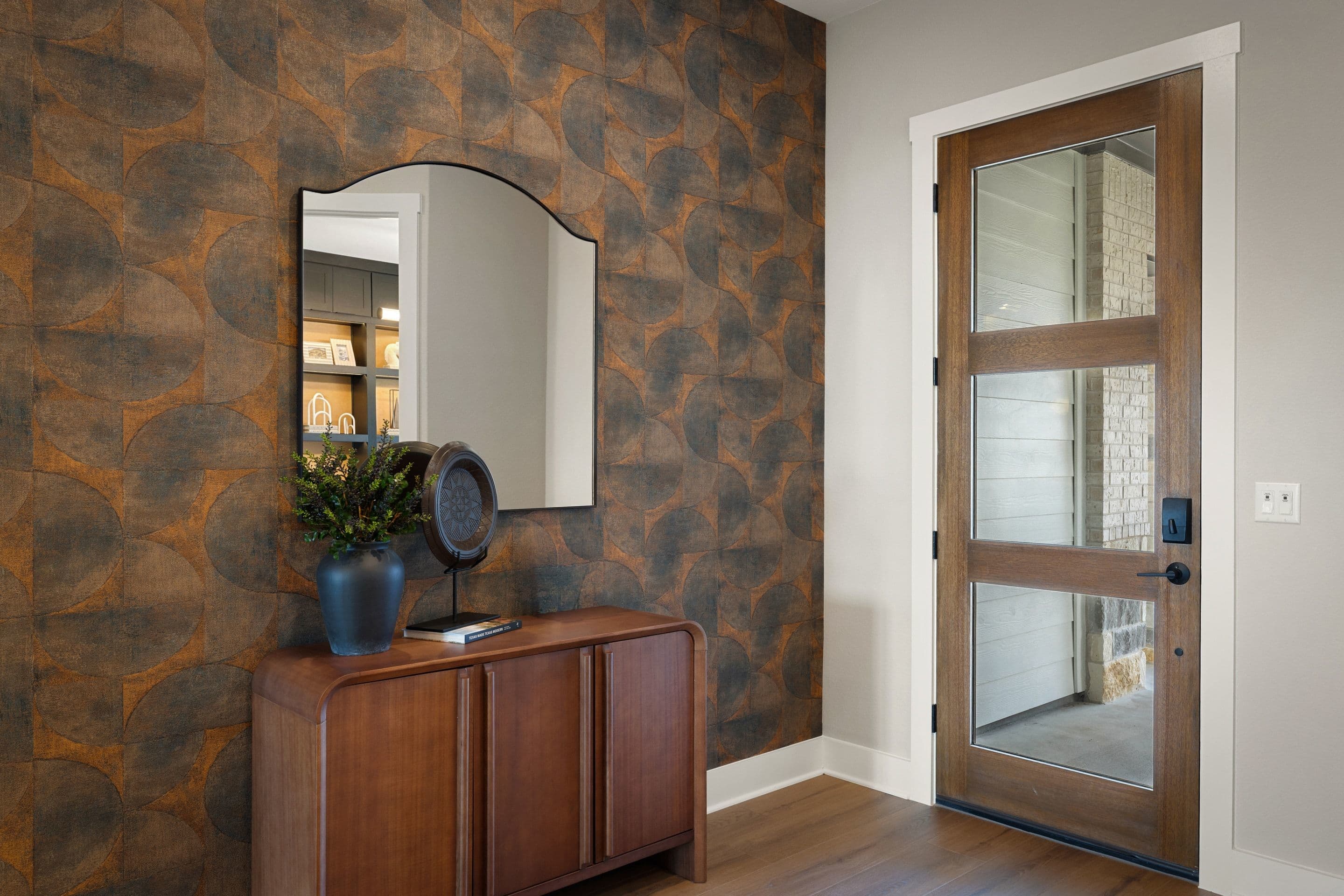 A wooden cabinet with a mirror and plant sits against a textured wall, while a glass-paneled door stands to the right.