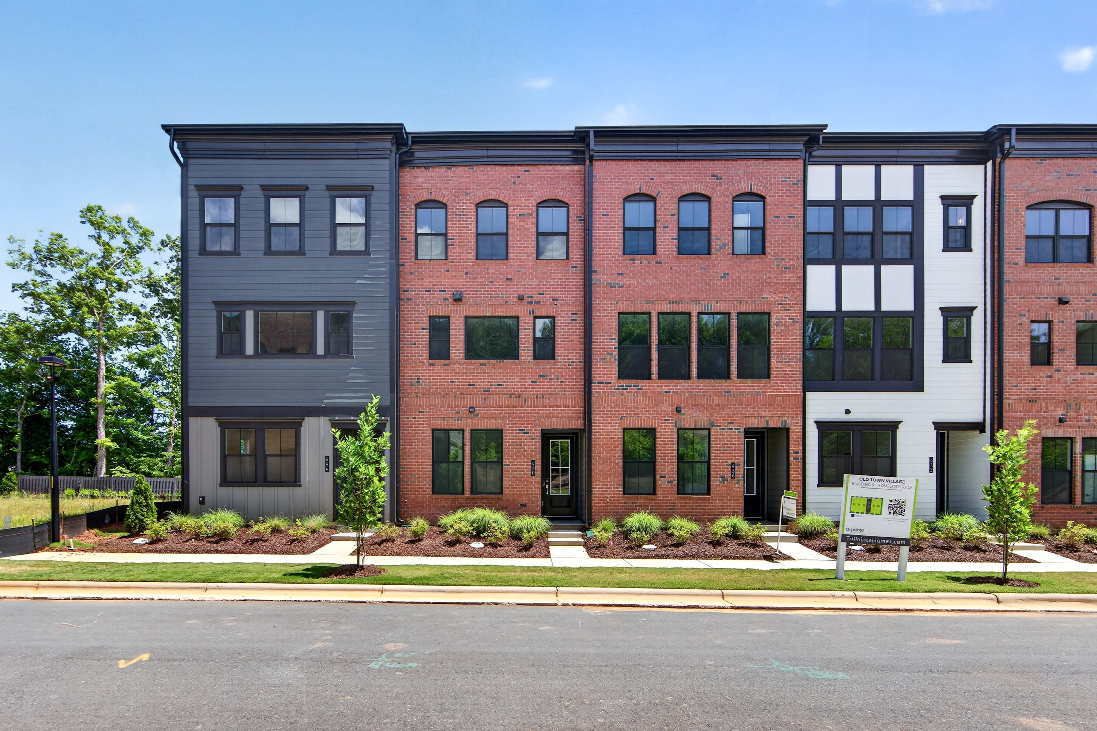 A row of modern townhouses with manicured landscaping in the foreground stands against a clear blue sky with scattered trees in the background.