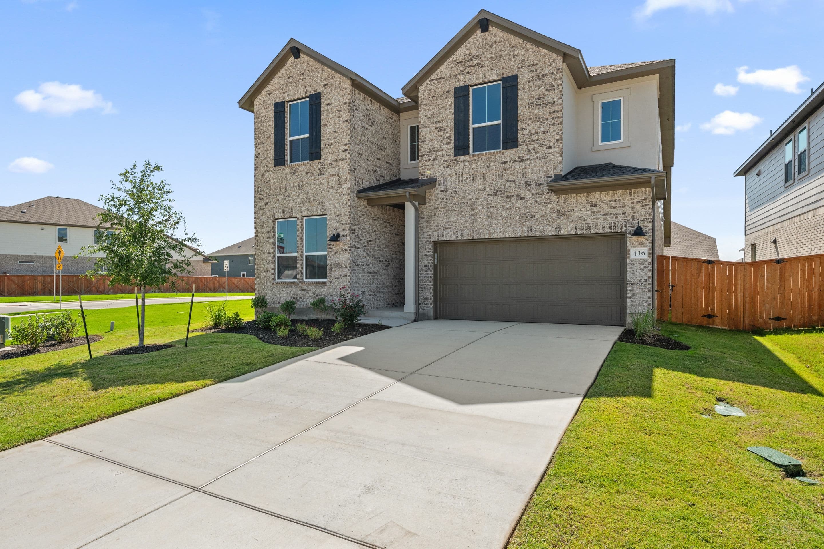 A modern brick house with a two-car garage sits on a manicured lawn, with a clear blue sky and neighboring houses in the background.