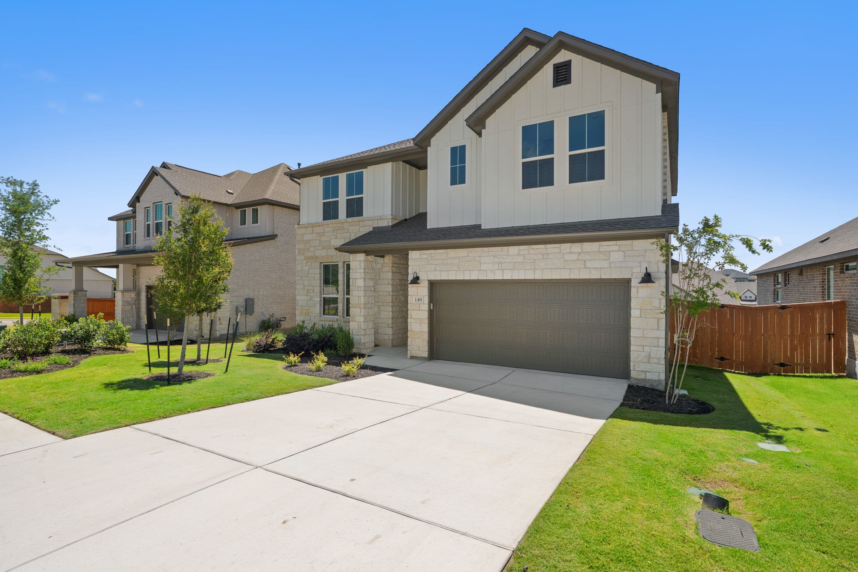 A modern two-story house with a light-colored stone and siding exterior sits on a manicured lawn with a concrete driveway in the foreground, while other houses and a wooden fence are visible in the background under a clear blue sky.