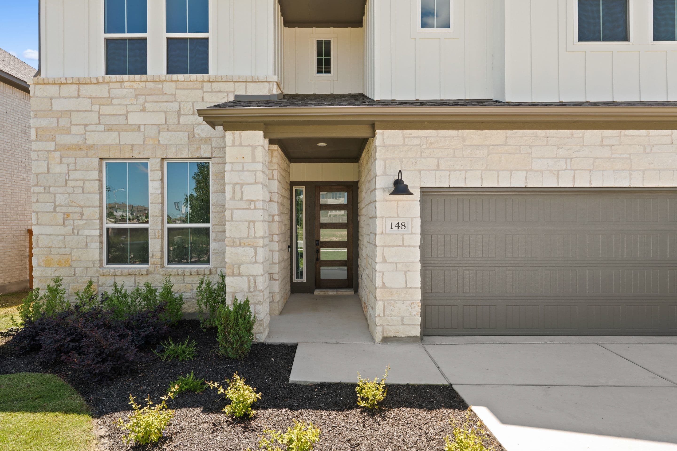 A modern house with a stone facade and a dark garage door is fronted by dark mulch and green bushes, with a clear blue sky visible above.