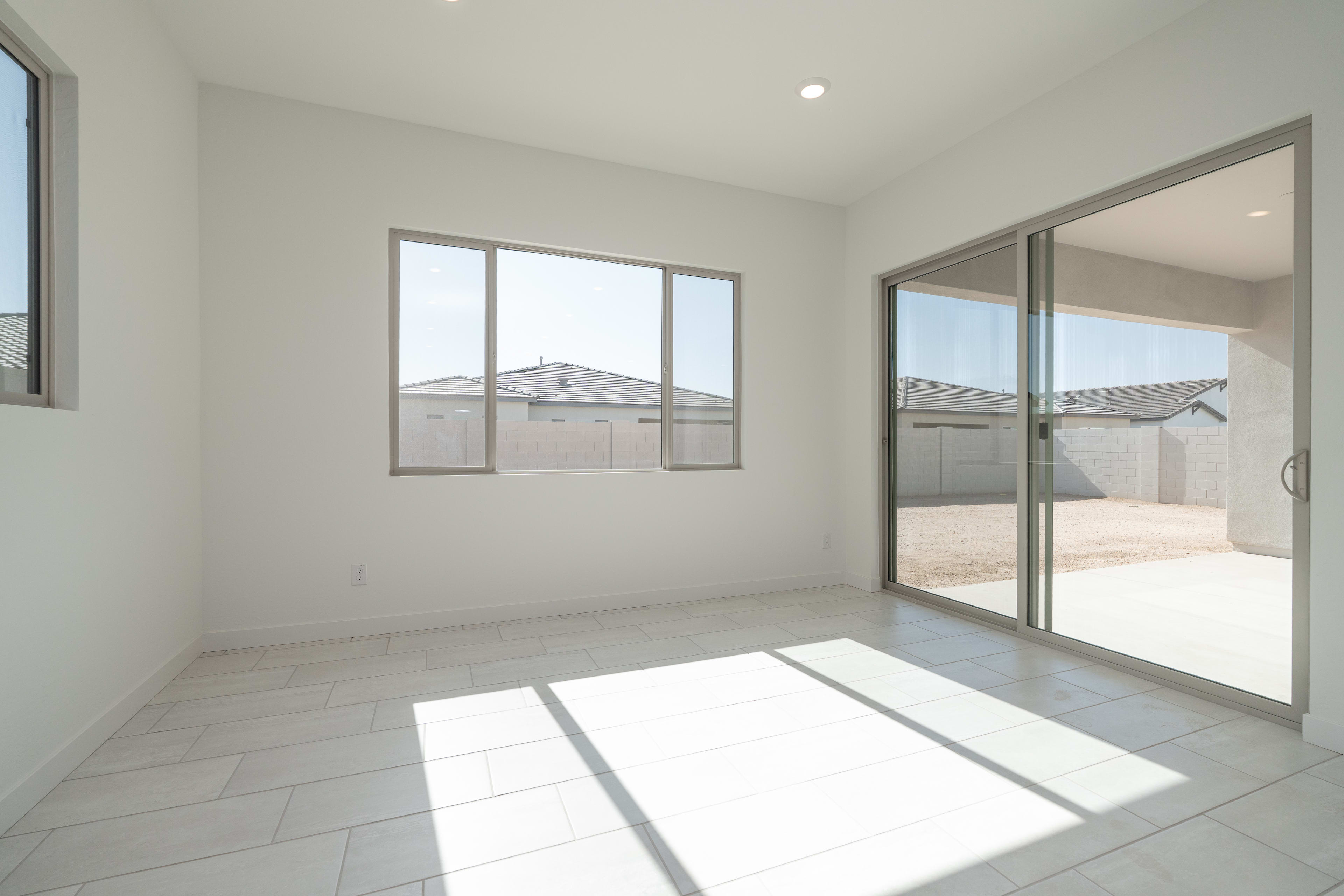 Sunlight streams across the white tiled floor of an empty room with a large window and sliding glass doors that open to a backyard with houses and a garage.