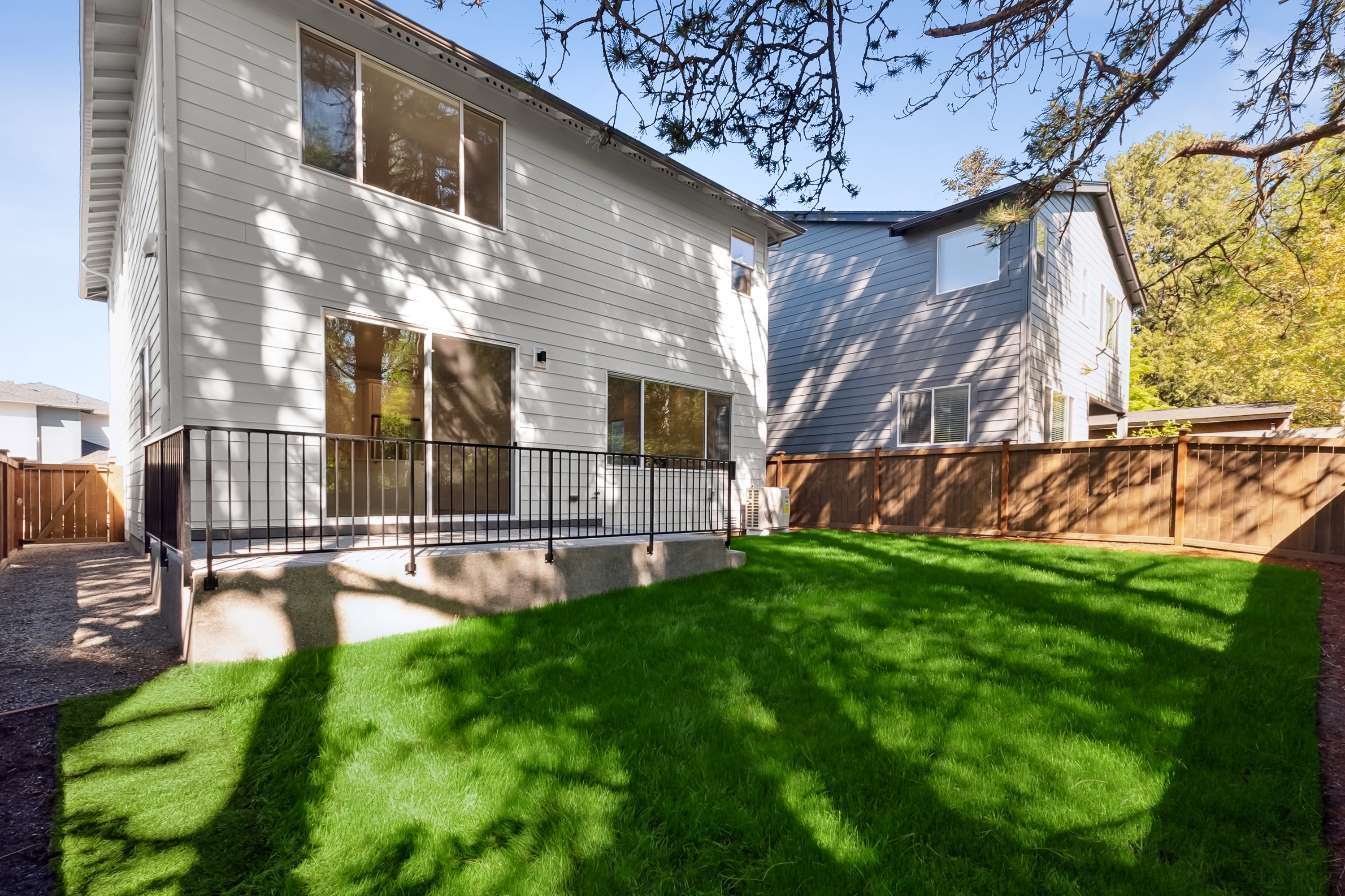 A bright green lawn with dappled sunlight covers the foreground, leading to a white two-story house with sliding glass doors and a black railing in the midground, while a neighboring house and wooden fence form the background.