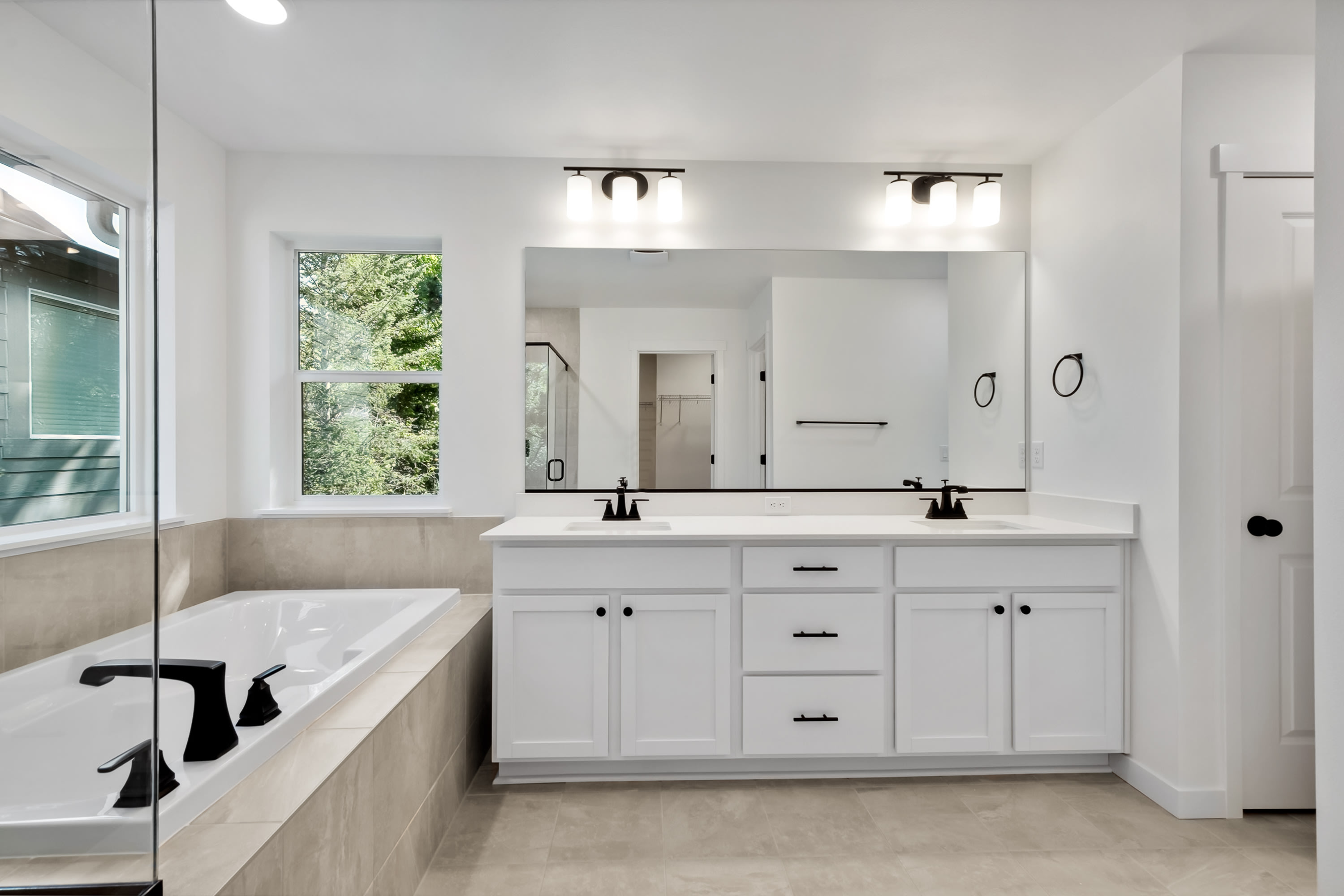 A modern bathroom features a white soaking tub in the foreground and a double vanity with a large mirror reflecting a closet in the background.